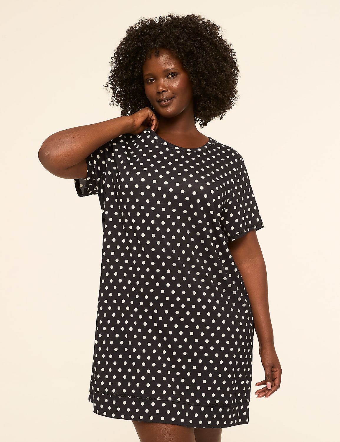 A woman with dark, curly hair stands in a black and white polka dot dress against a plain, light-colored background. Product Image 1