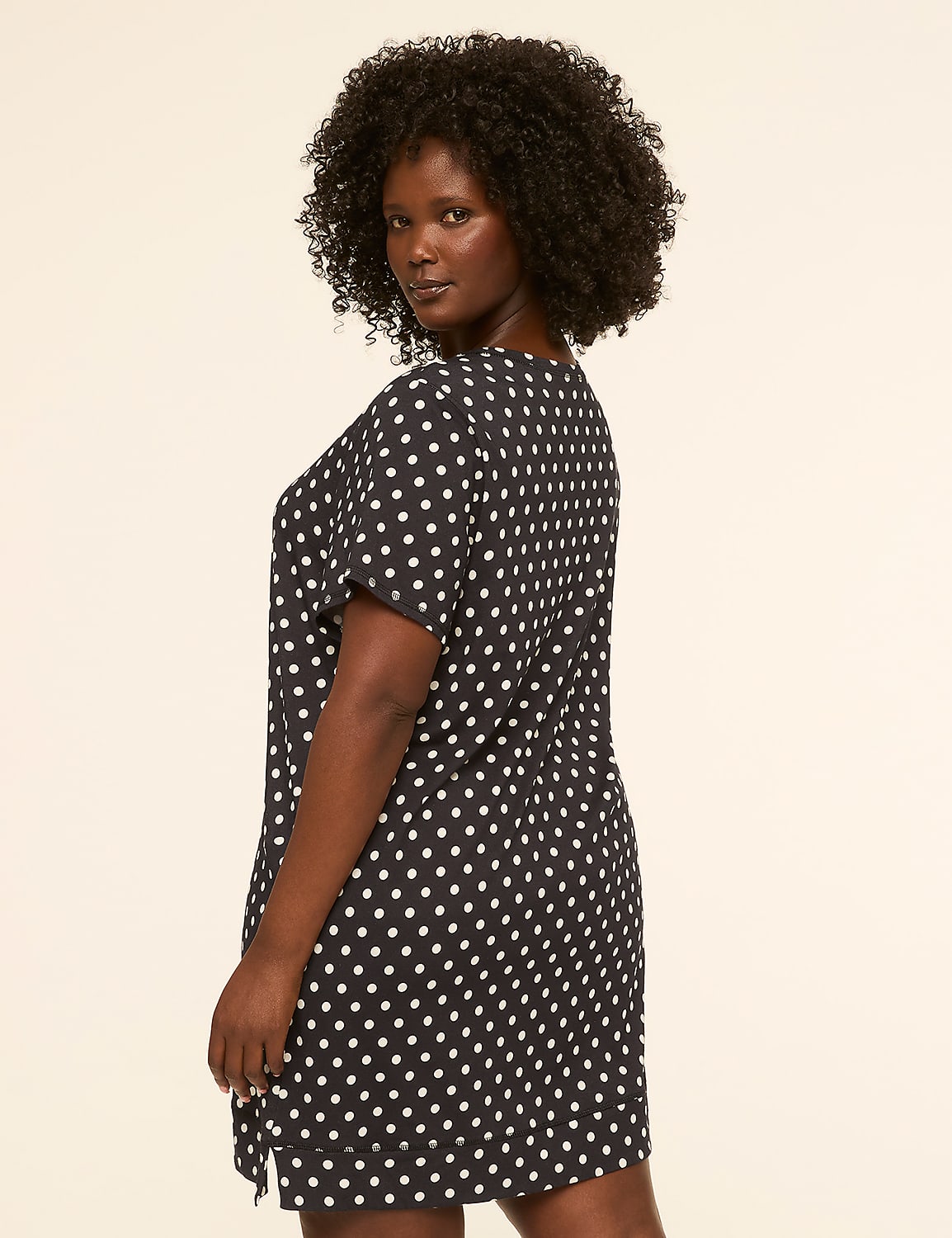 A woman with dark, curly hair poses in a dark, polka-dotted dress against a plain, light-colored background. Product Image 2