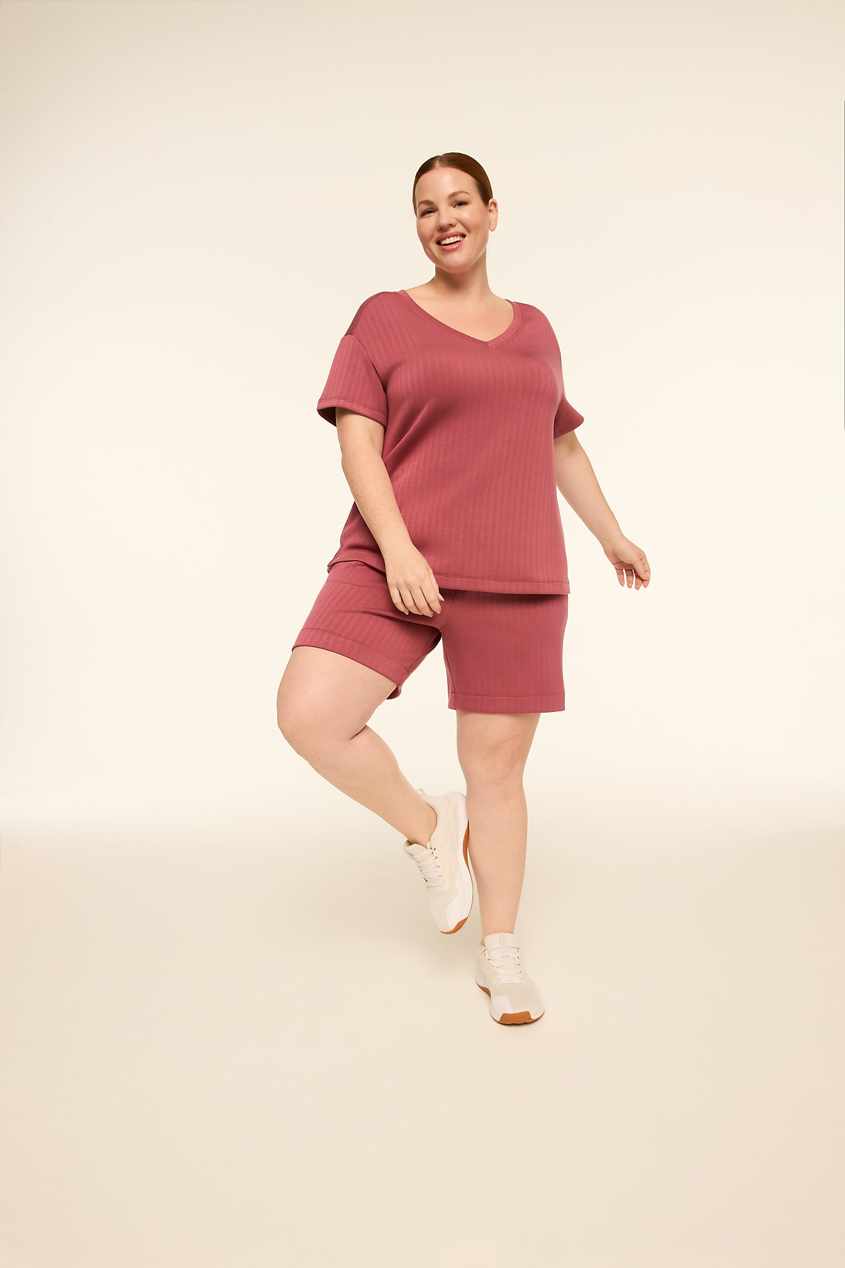 A smiling woman in a mauve loungewear set and white sneakers stands on a plain, light-colored studio floor against a soft, gradient background. Product Image 4