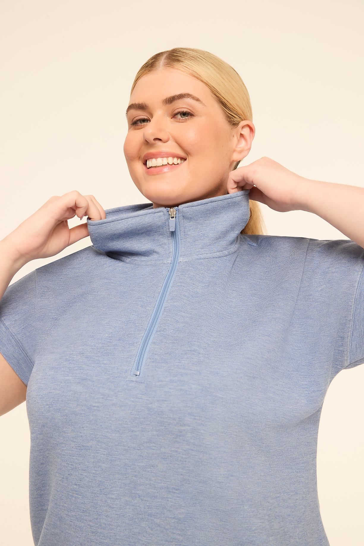 A smiling woman in a blue zip-neck top adjusts the collar against a plain, light background. Product Image 5