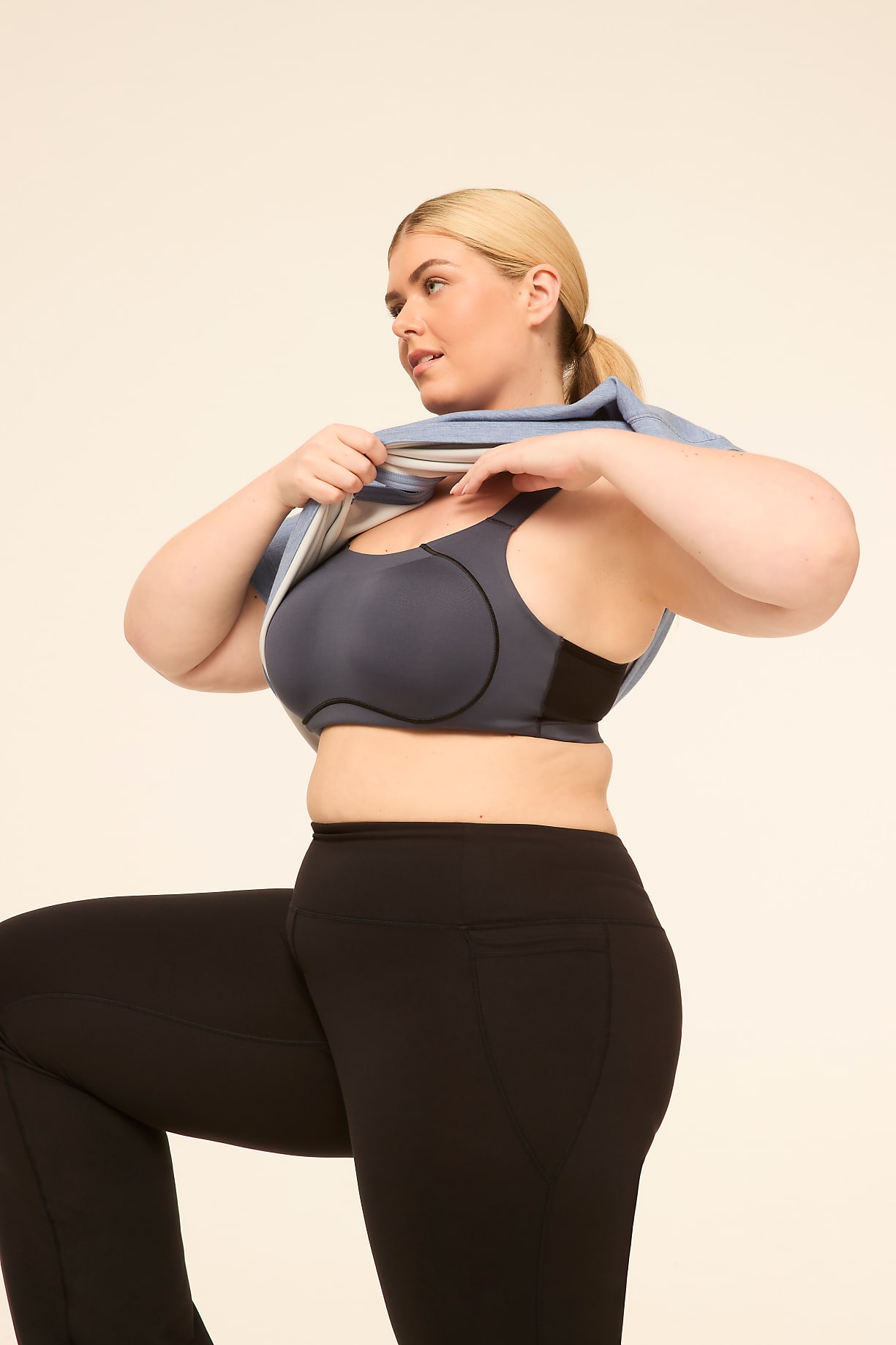 A woman in a sports bra and leggings pulls a blue jacket over her head against a plain background. Product Image 6
