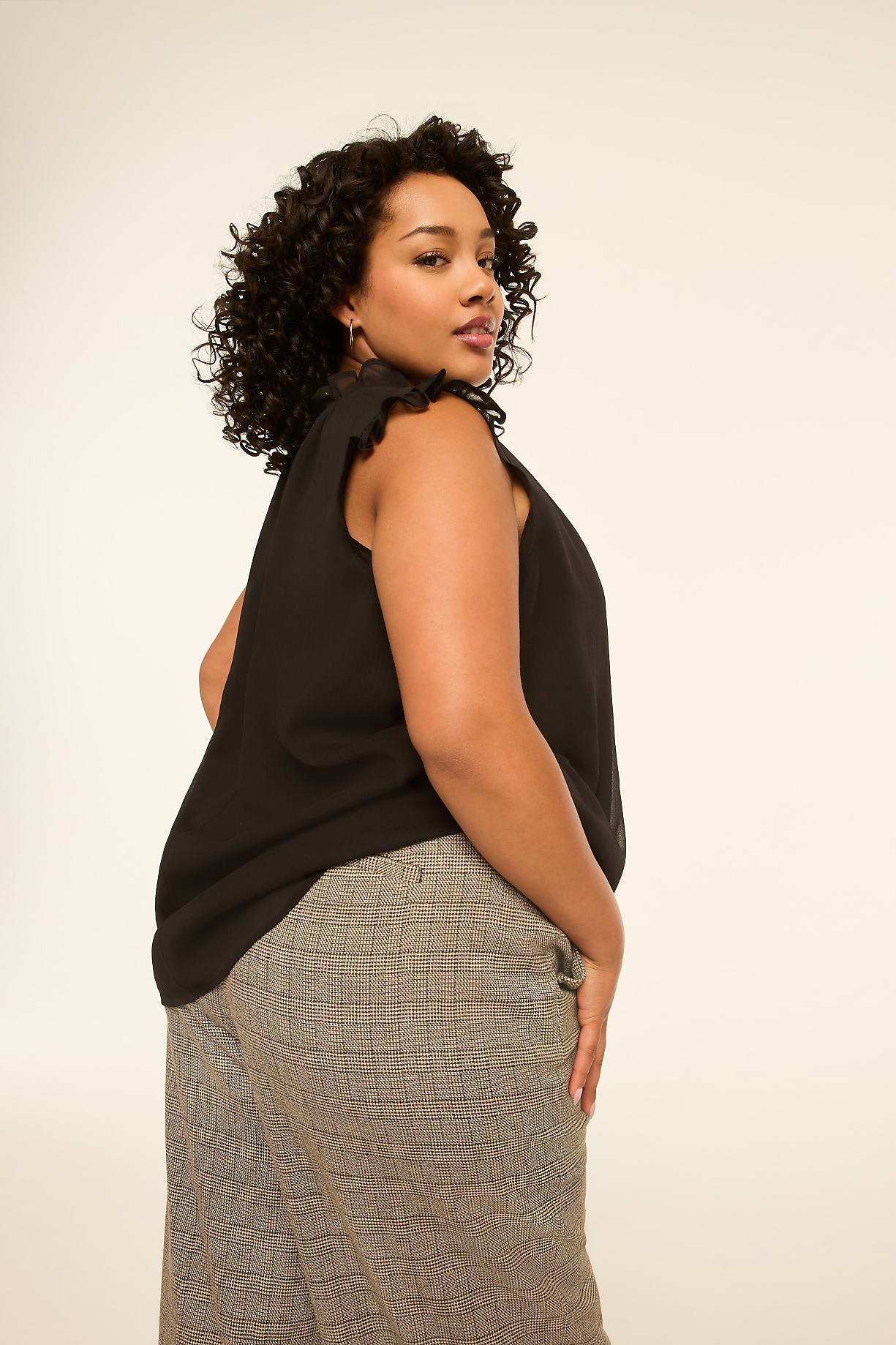 A woman with curly dark hair poses in a black top and patterned pants against a plain, light background. Product Image 4