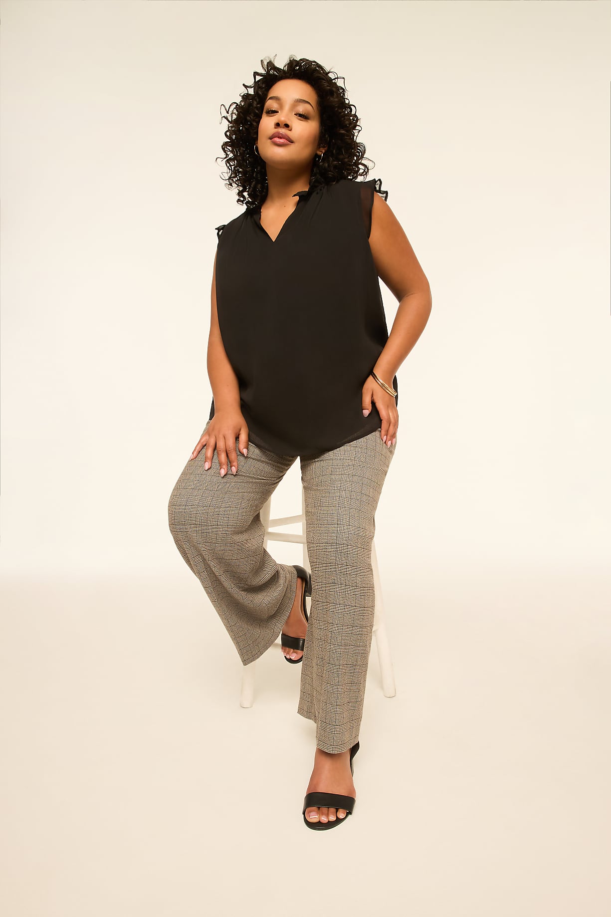 A woman with curly dark hair, wearing a black V-neck top and patterned pants, sits on a white stool against a plain, light-colored background. Product Image 5