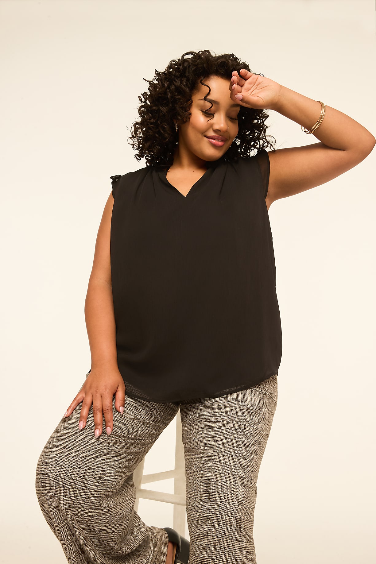 A woman with curly dark hair and a black top poses with her hand on her forehead against a plain, light background. Product Image 6