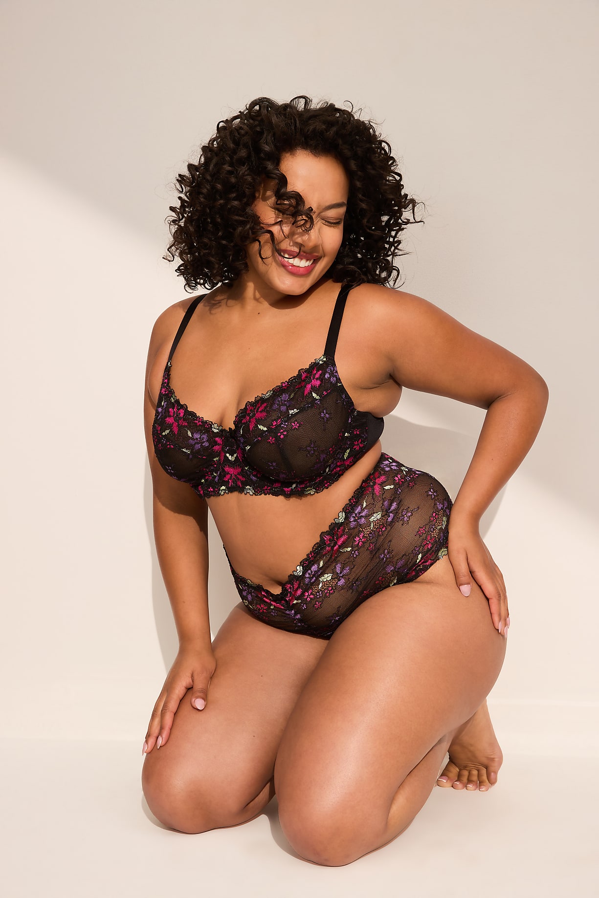 A smiling woman with curly hair kneels in a black floral bra and underwear against a plain, light-colored background. Product Image 4