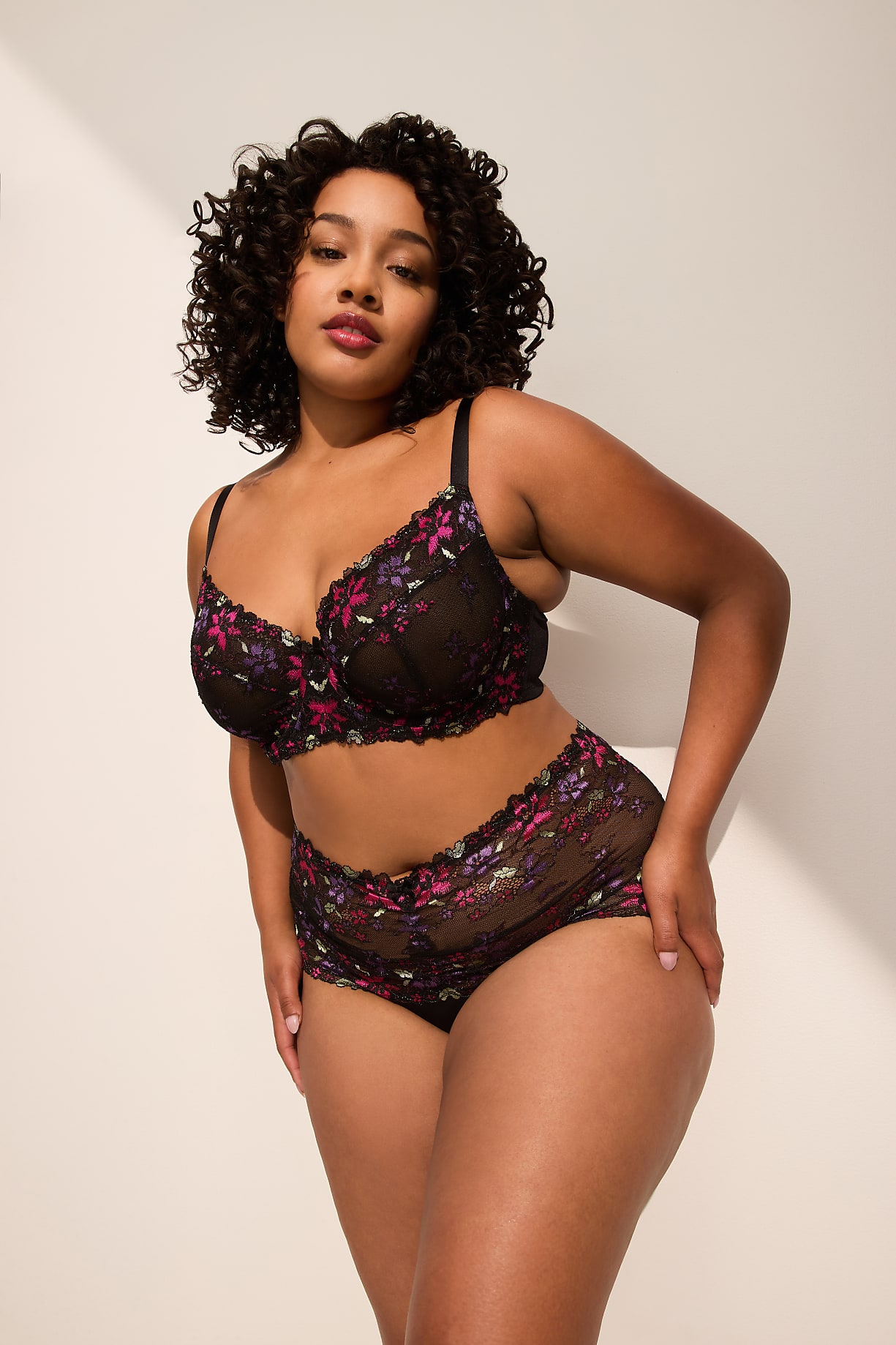 A woman with curly dark hair poses in a floral lace bra and matching underwear against a plain white background with a shadow on the wall. Product Image 5