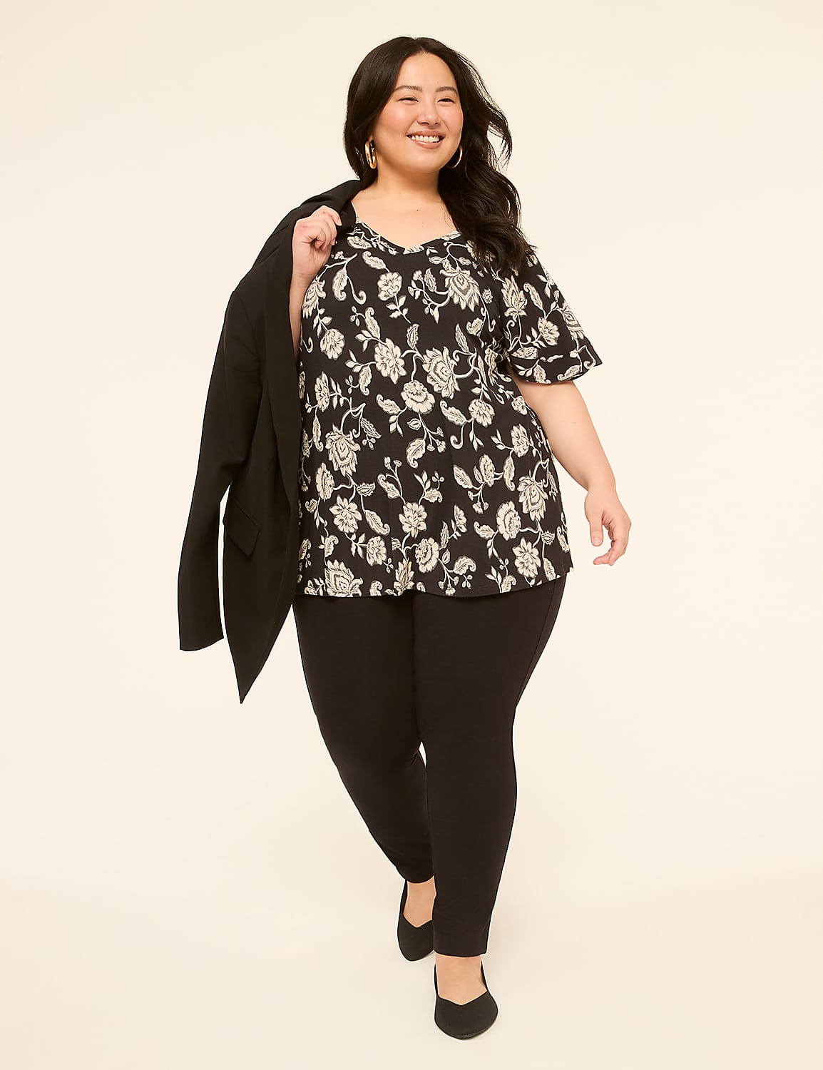 A smiling woman in a floral top and black pants walks forward with a blazer draped over her shoulder against a plain, light-colored background. Product Image 3