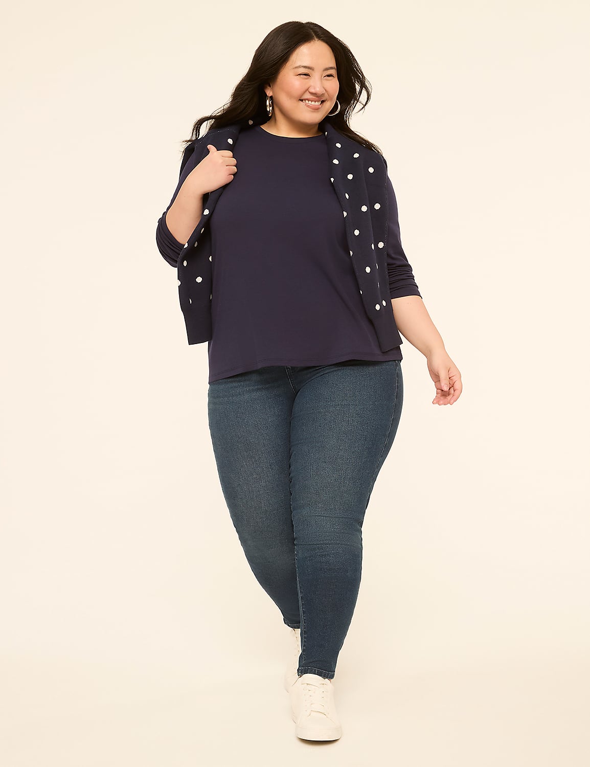 A smiling woman in a navy shirt, polka dot cardigan, and jeans walks against a plain, light-colored background. Product Image 3