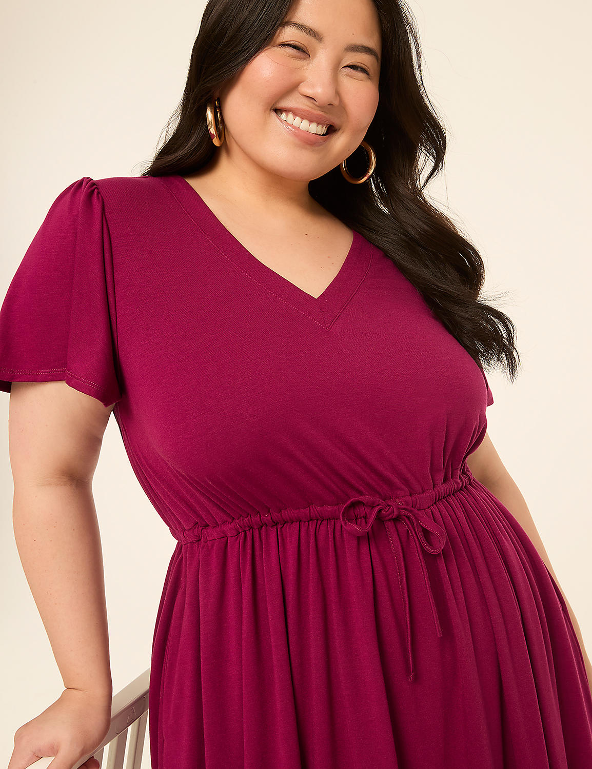 A smiling woman in a magenta dress with flutter sleeves and a drawstring waist is leaning on a chair against a plain, light-colored background. Product Image 3