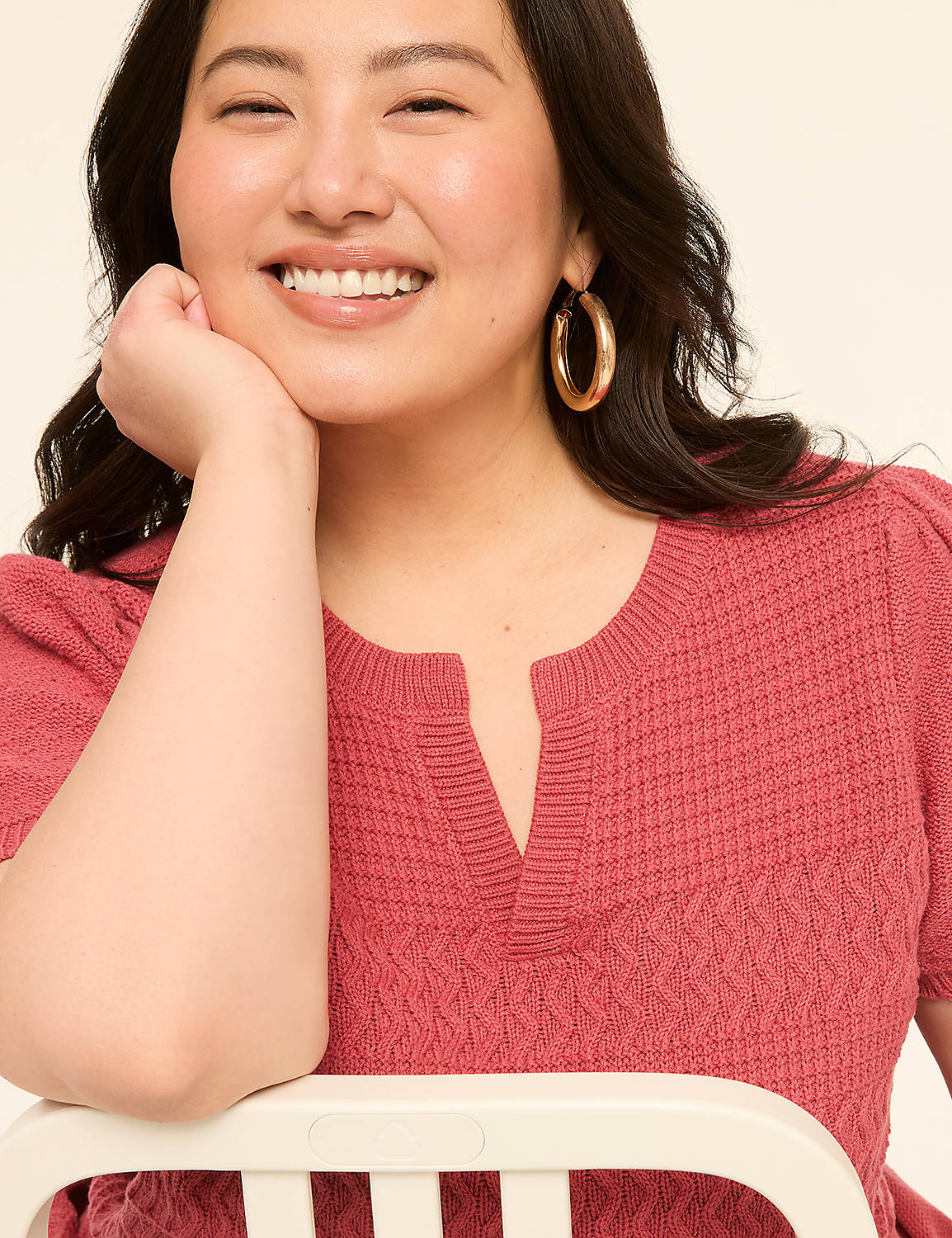 A smiling woman with dark hair and hoop earrings rests her chin on her hand, wearing a textured pink top against a plain cream background. Product Image 4