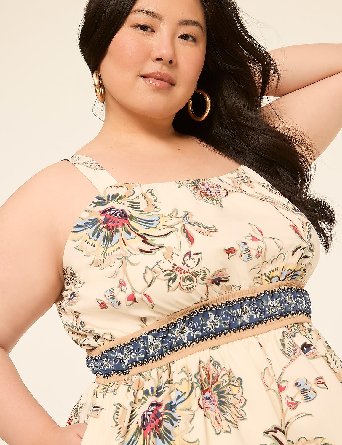 A woman with dark hair and hoop earrings smiles as she poses in a cream-colored floral dress with a blue and tan patterned waistband against a plain background. Product Image 3