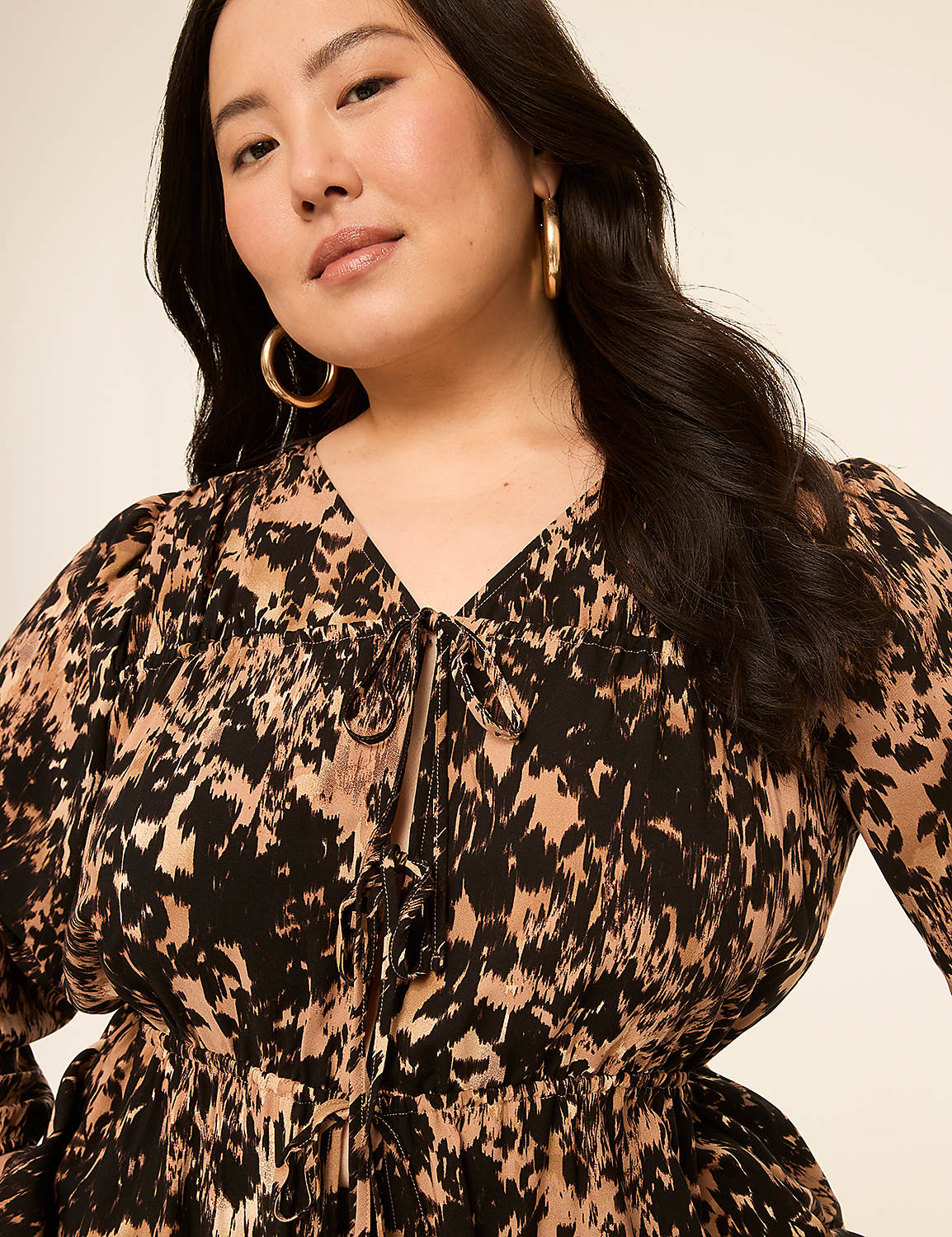A woman with dark hair and hoop earrings smiles gently in the foreground, wearing a black and tan abstract print dress, against a plain, light-colored background. Product Image 4