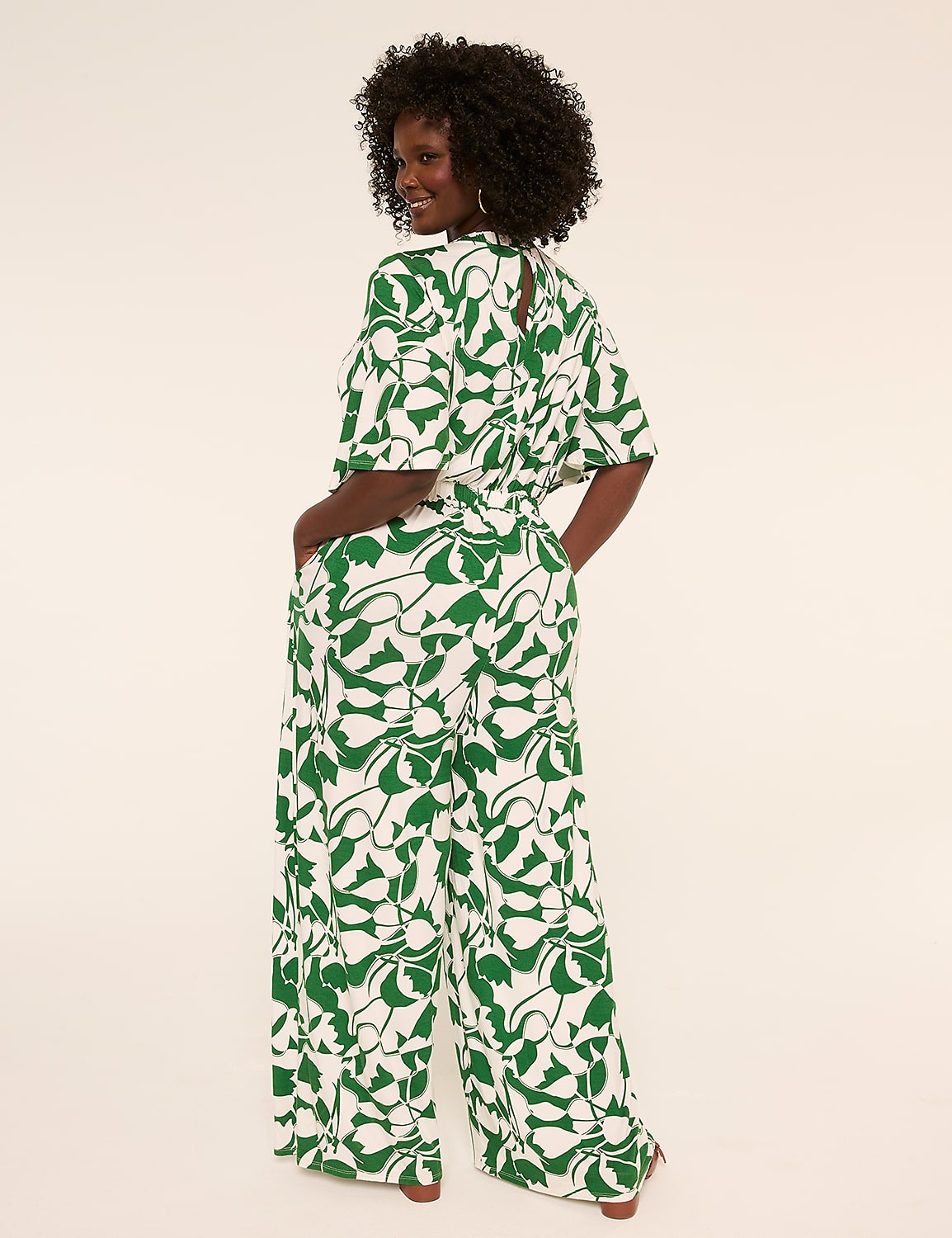 A woman in a green and white patterned jumpsuit stands with her hands in her pockets against a plain, light-colored background. Product Image 2
