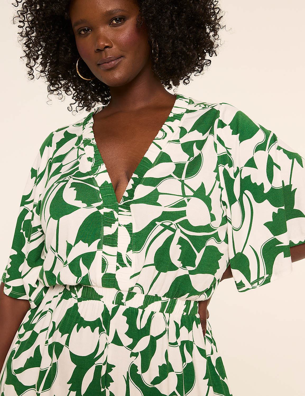 A woman with curly dark hair and hoop earrings smiles gently, wearing a white and green floral print dress against a plain, light background. Product Image 3