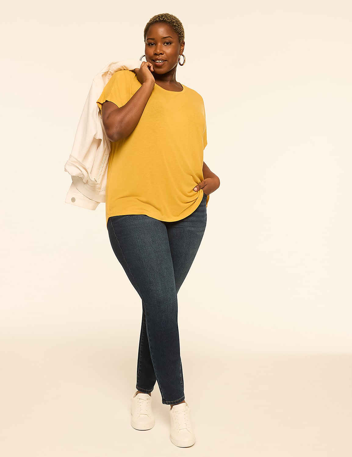 A woman in a mustard yellow t-shirt and dark wash jeans stands against a plain, light-colored background with a white jacket draped over her shoulder. Product Image 3