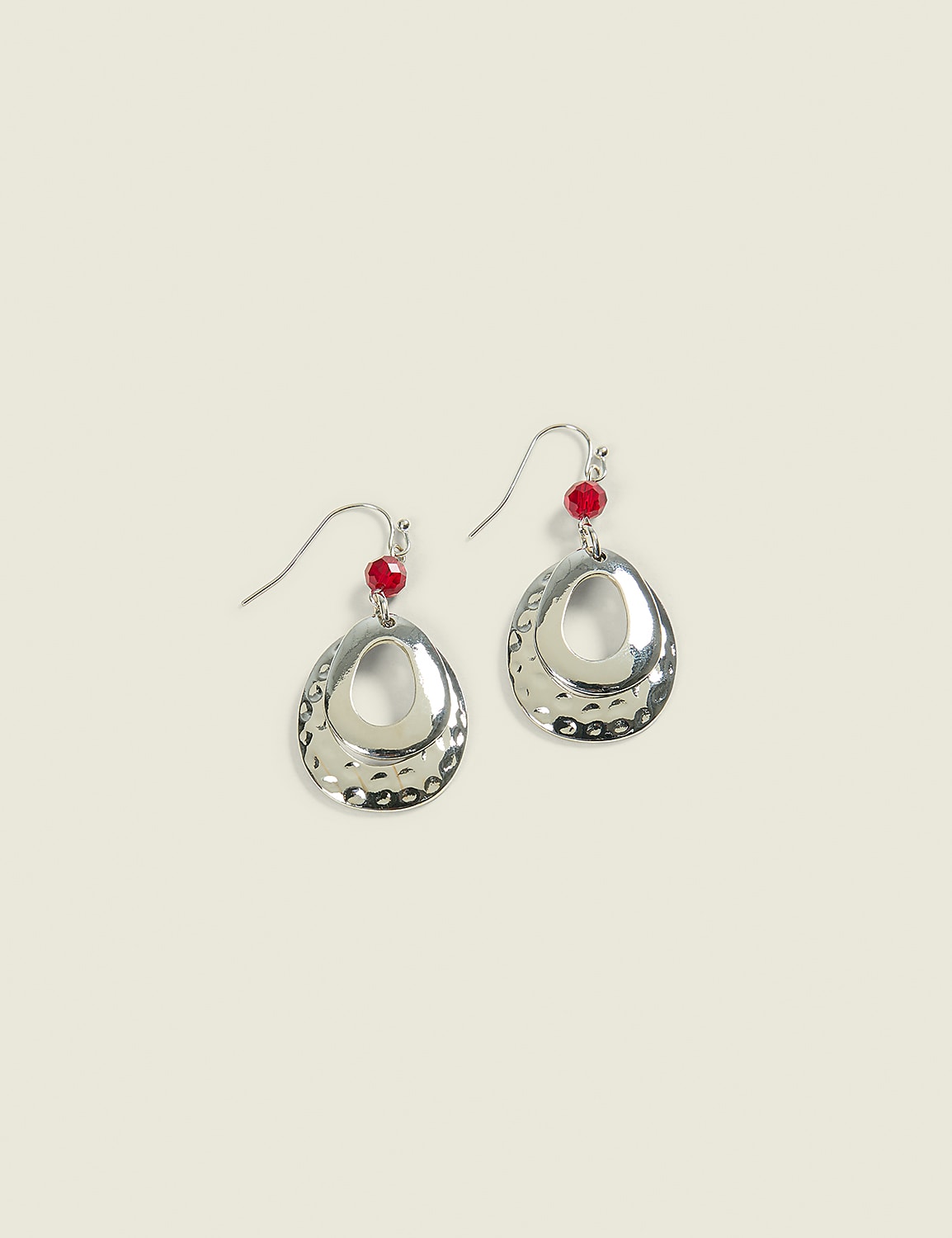A pair of silver, hammered oval earrings with red beads hang against a pale, textured background. Product Image 1