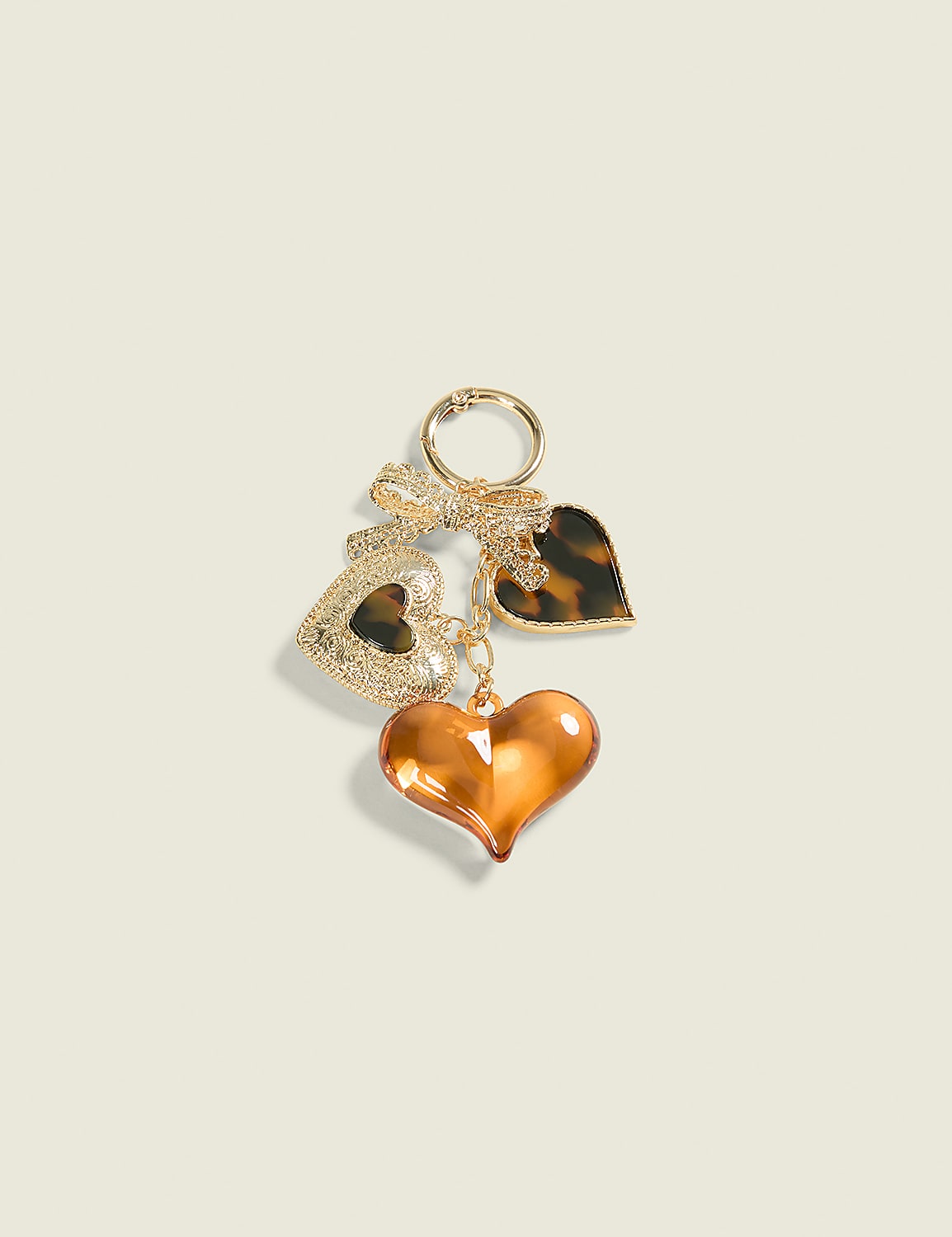 A gold keychain with three heart-shaped charms, including one amber-colored, one tortoiseshell, and one glittery gold, hangs against a plain, light beige background. Product Image 1