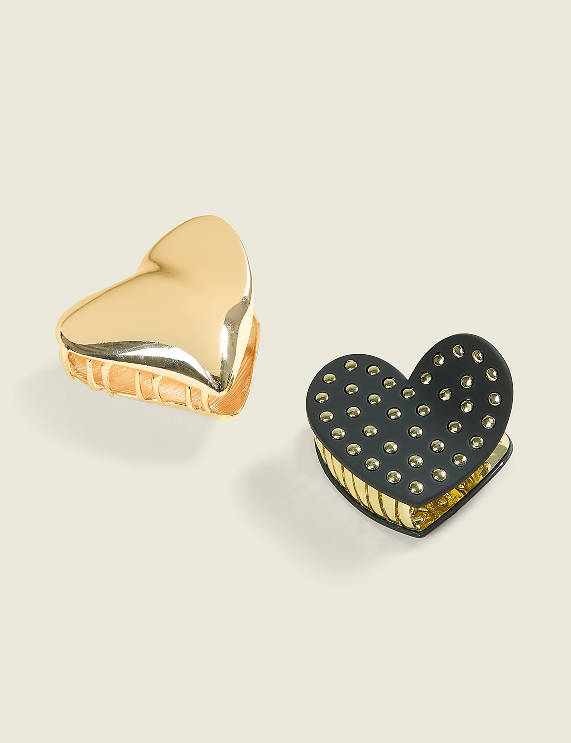 Two heart-shaped hair clips, one gold and one black with gold studs, rest on a pale, neutral background. Product Image 1
