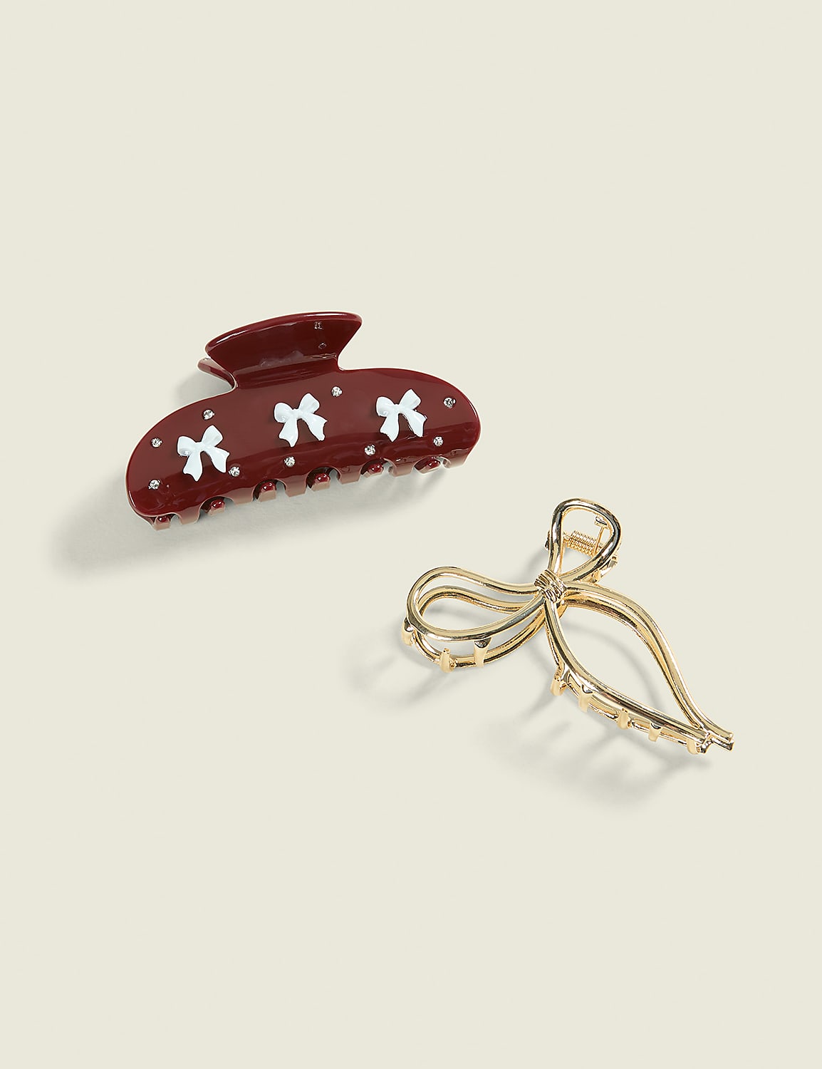 Two decorative hair clips, one brown with white bows and the other gold with a ribbon design, rest on a plain, light-colored background. Product Image 1