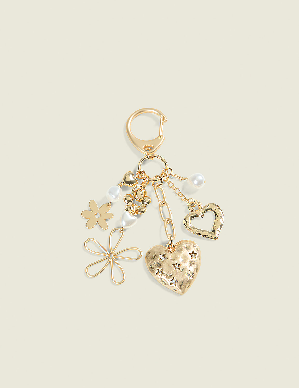 A gold keychain with various charms, including a textured heart, a wire flower, a pearl, and a small flower, hangs against a pale yellow background. Product Image 1
