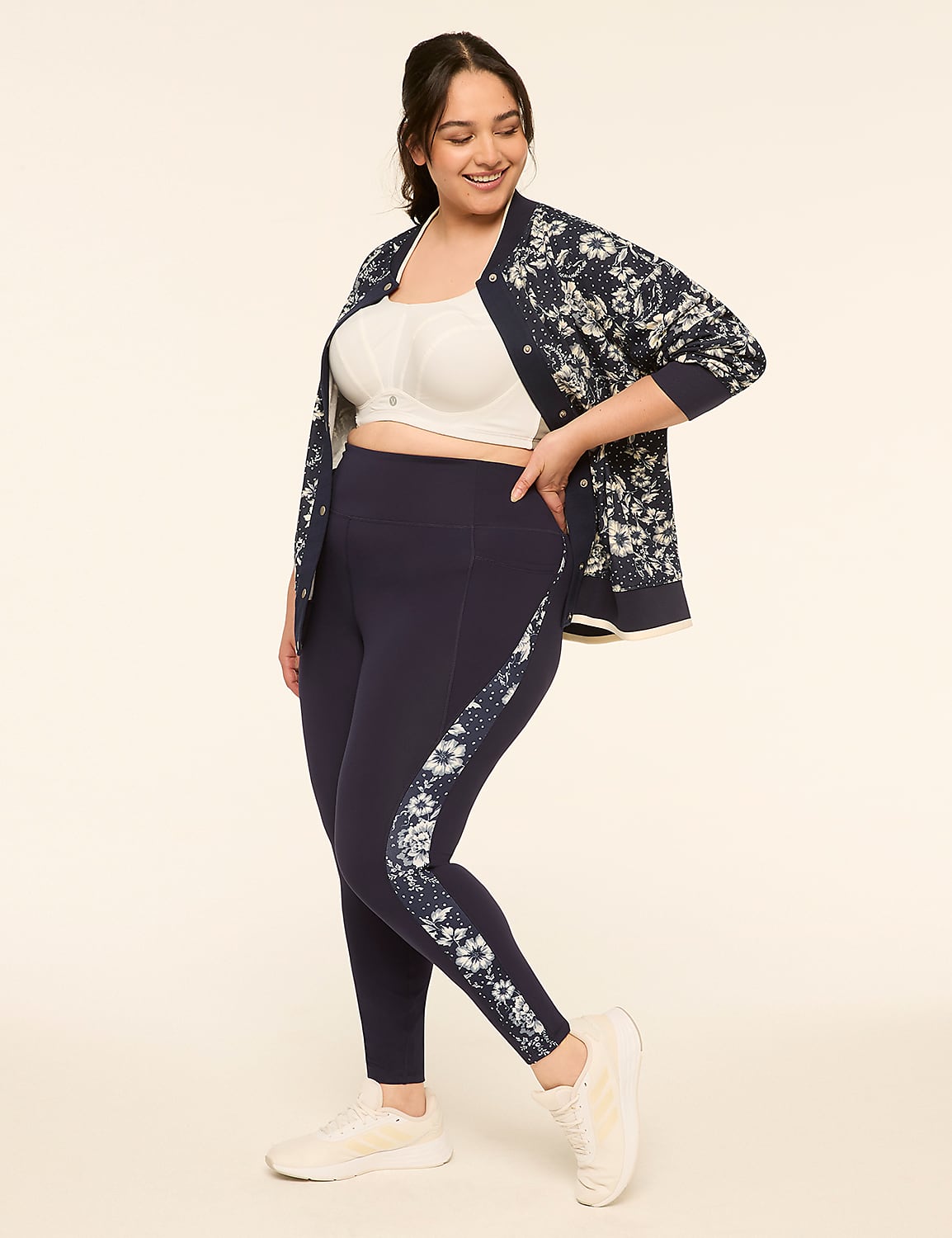 A woman in a white sports bra, navy floral bomber jacket, and navy leggings with a floral stripe stands against a plain, light-colored background. Product Image 3
