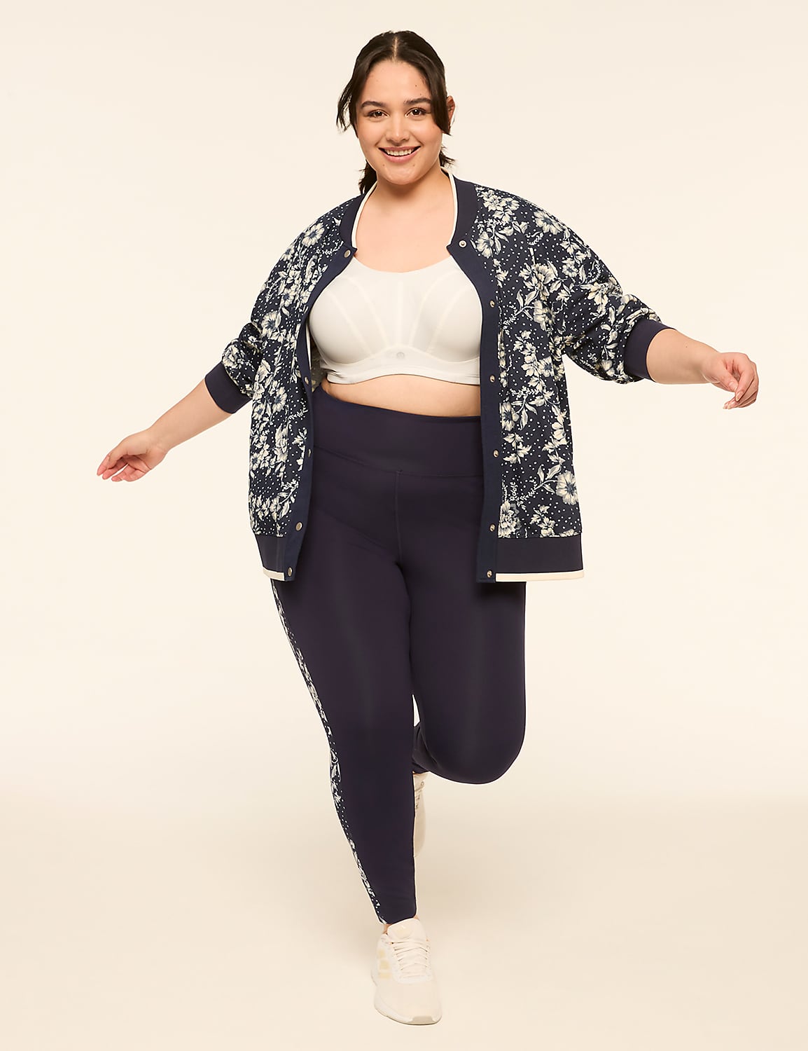 A smiling woman in a floral bomber jacket, white sports bra, and navy leggings poses against a plain cream background. Product Image 3