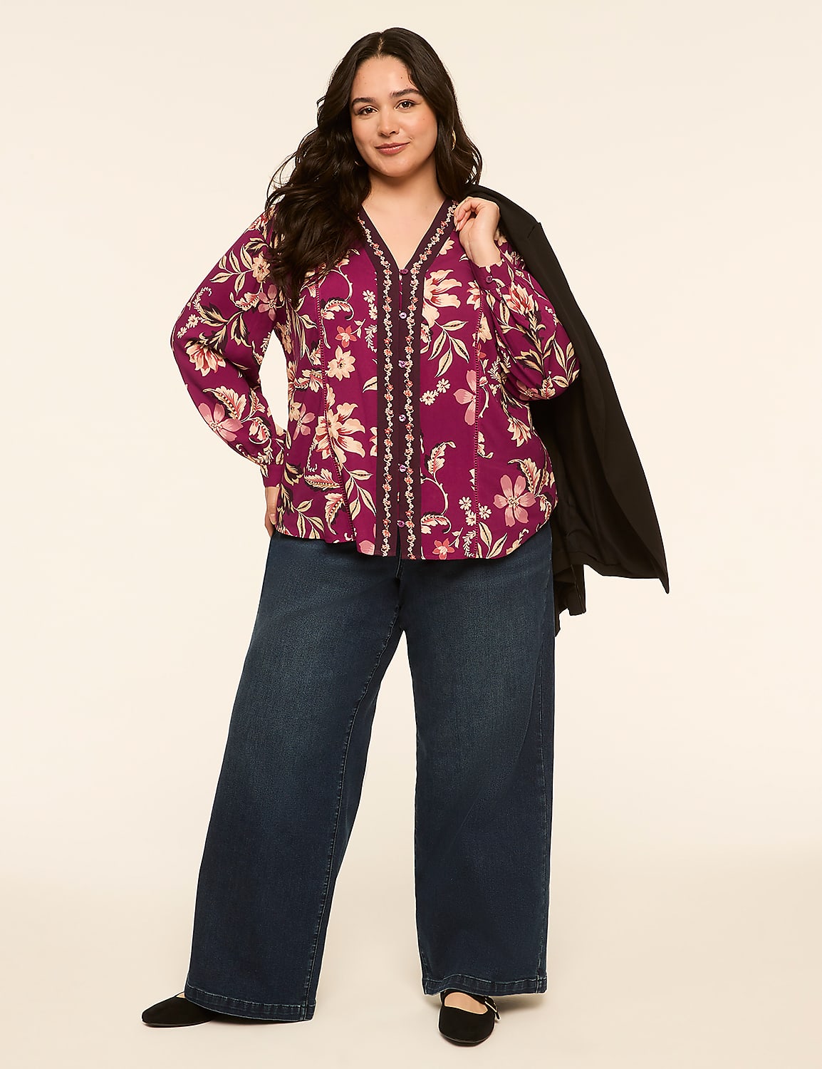 A woman in a floral blouse and wide-leg jeans stands against a plain, light-colored background, with a black jacket draped over her shoulder. Product Image 3