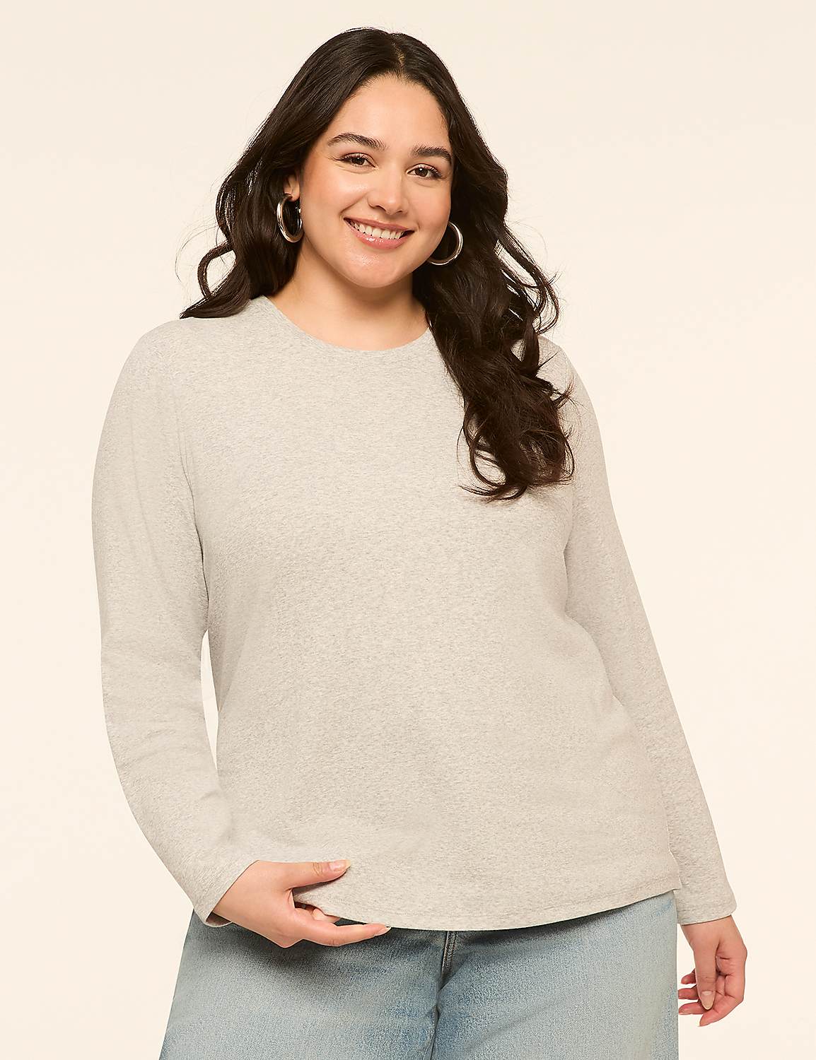 A smiling woman with dark hair and hoop earrings in a light gray long-sleeved shirt and blue jeans stands against a plain, light beige background. Product Image 1