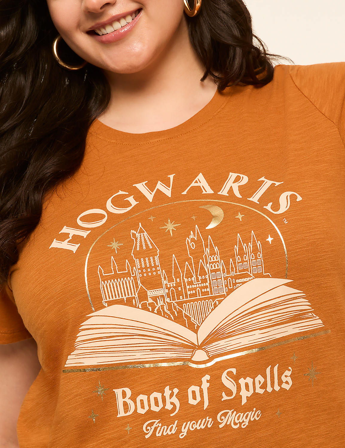 A woman smiles as she wears a burnt orange t-shirt with a gold foil print of Hogwarts castle on an open book, set against a plain background. Product Image 4