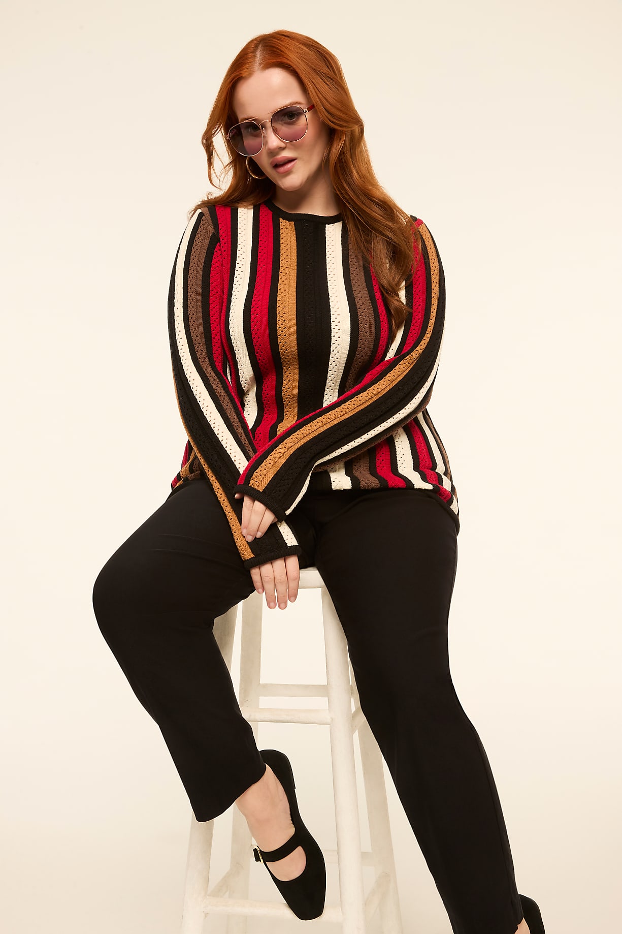 A woman with red hair sits on a white stool in the foreground, wearing a striped sweater and black pants, against a plain, light-colored background. Product Image 5