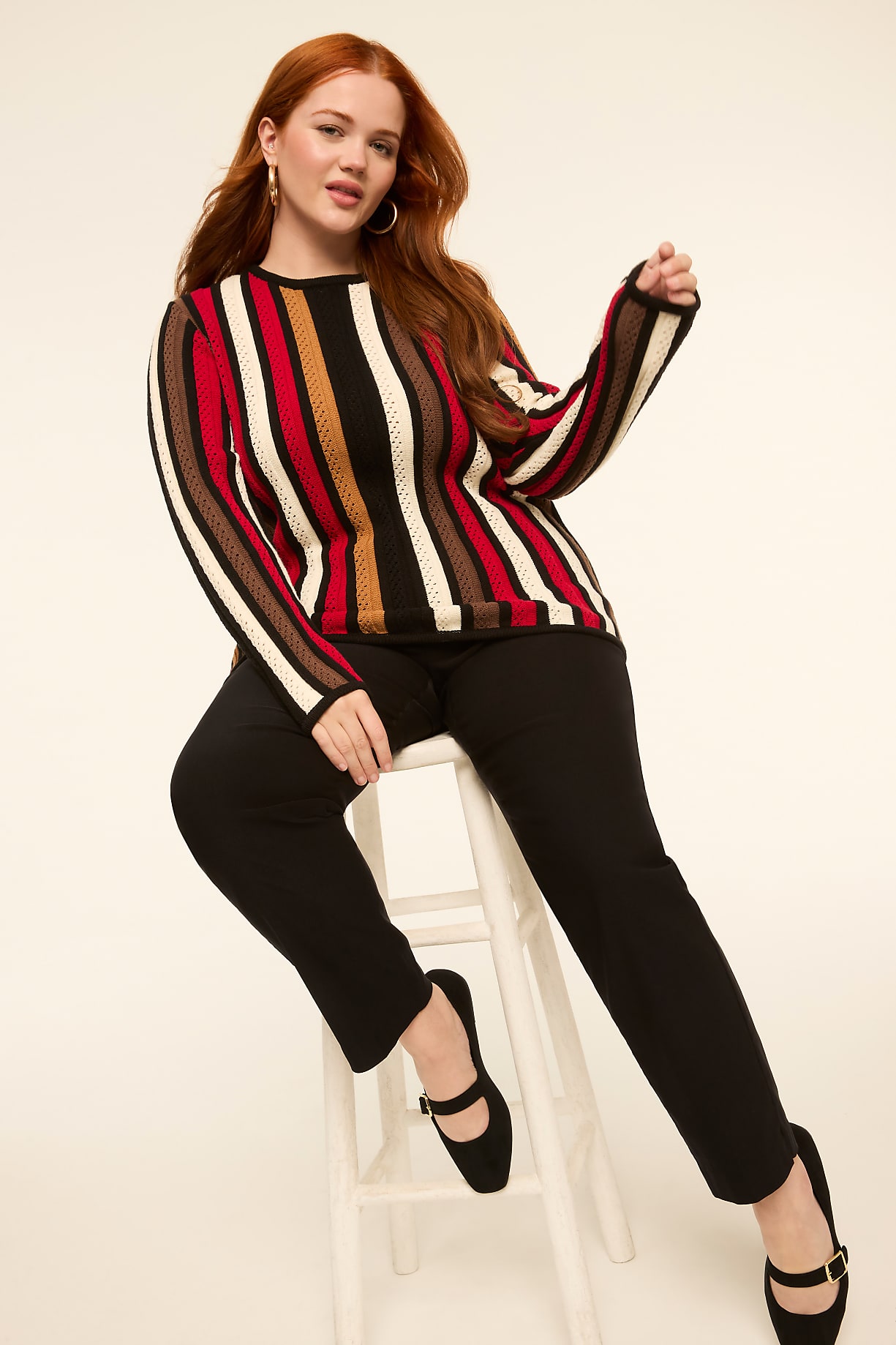 A woman with red hair sits on a white stool in the foreground, wearing a striped sweater and black pants, against a plain, light-colored background. Product Image 6
