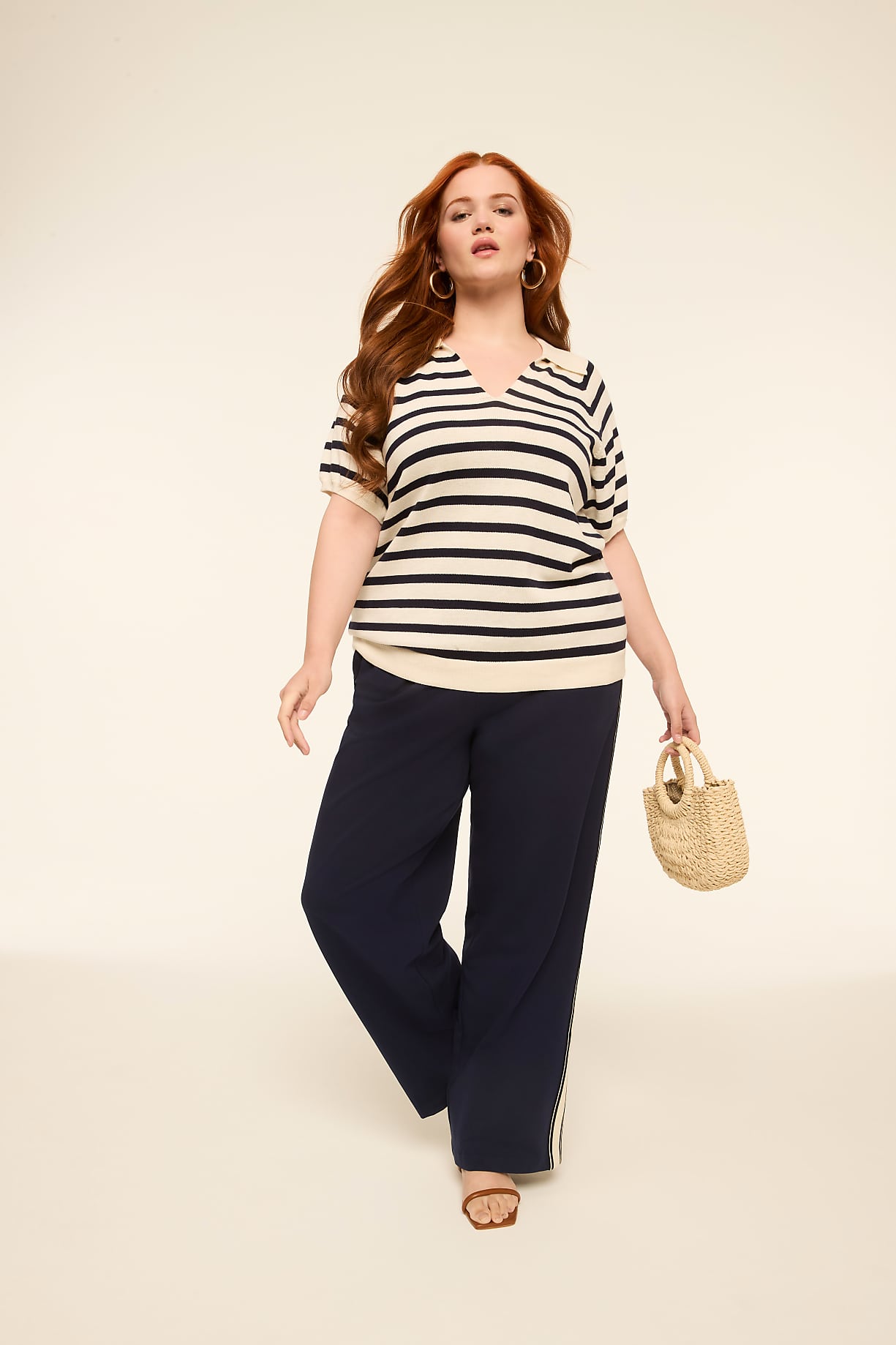 A woman with red hair wearing a striped shirt and dark pants walks forward holding a straw bag against a plain, light background. Product Image 3
