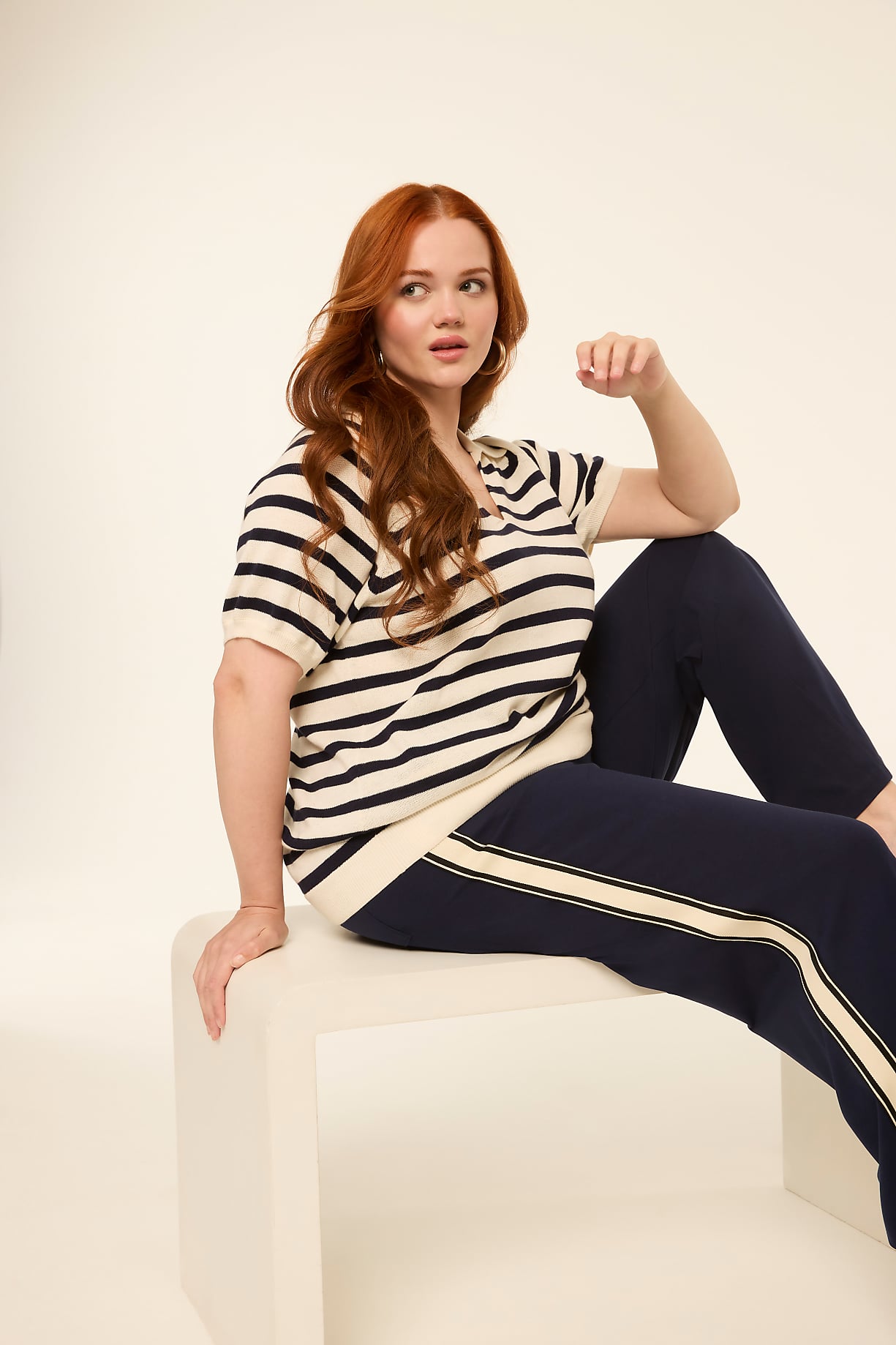 A woman with red hair sits on a white cube in the foreground, wearing a striped shirt and navy pants, against a plain, light-colored background. Product Image 7