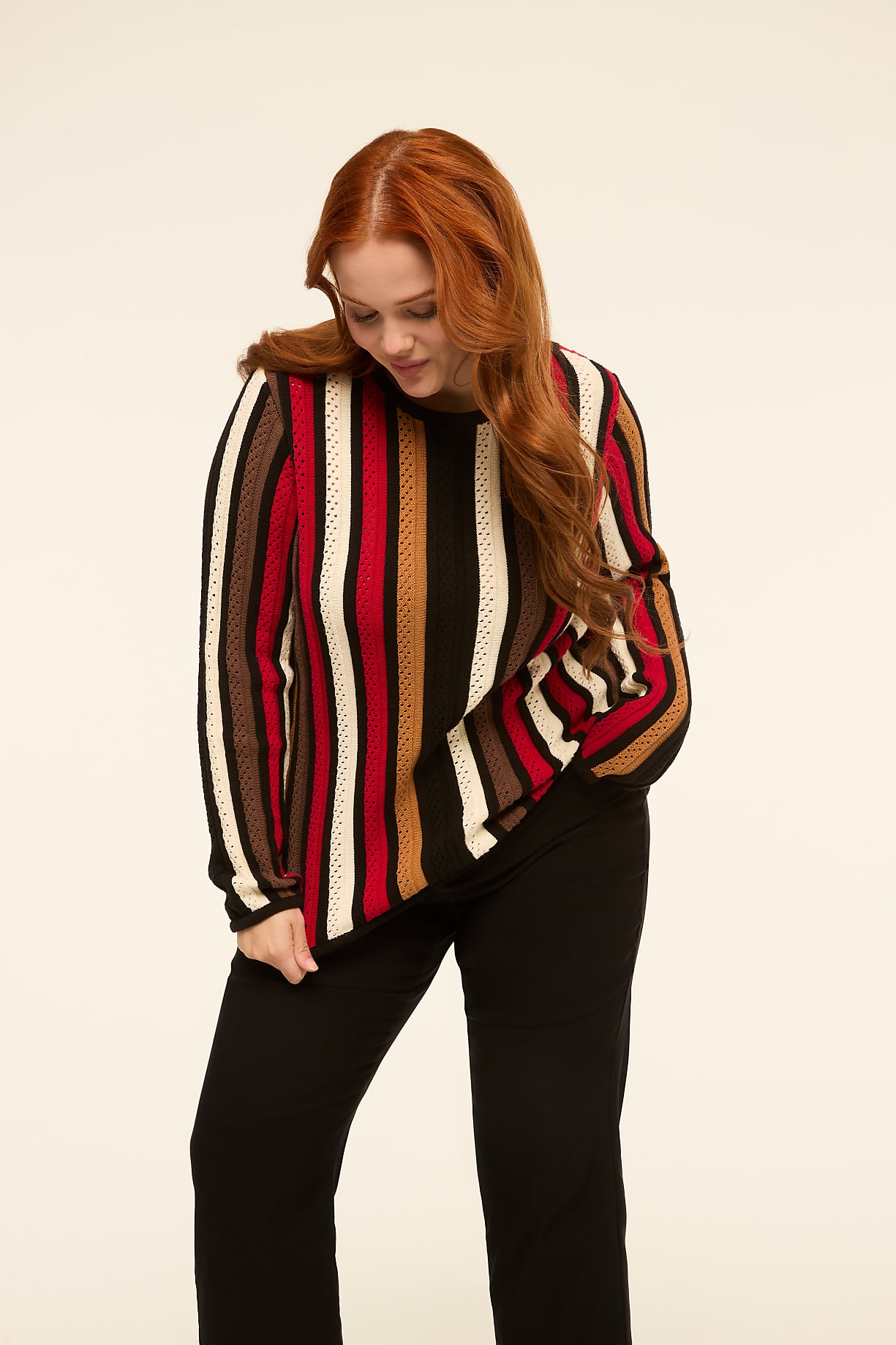A woman with long red hair stands in the foreground wearing a striped sweater and black pants, against a plain, light-colored background. Product Image 7
