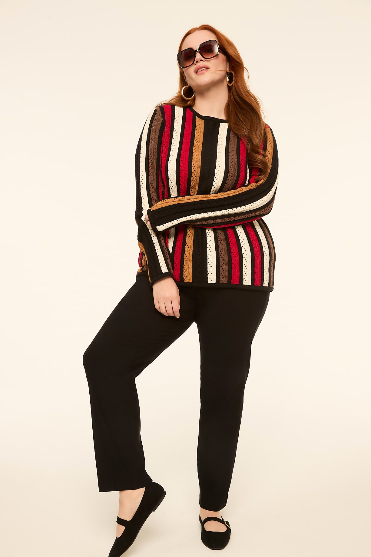 A woman with red hair and sunglasses poses in a striped sweater and black pants against a plain, light-colored background. Product Image 8