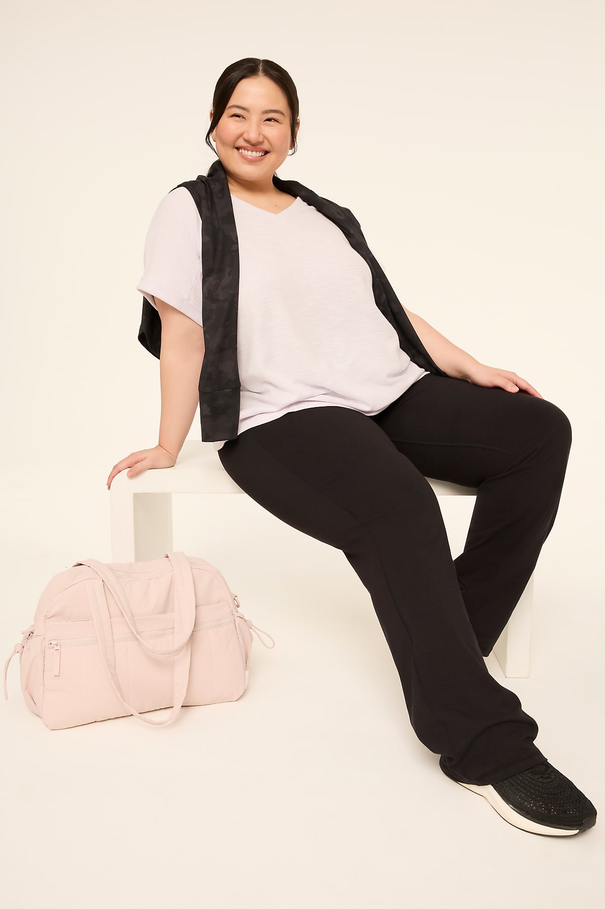 A smiling woman in athletic wear sits next to a pink duffel bag against a plain, light-colored background. Product Image 5