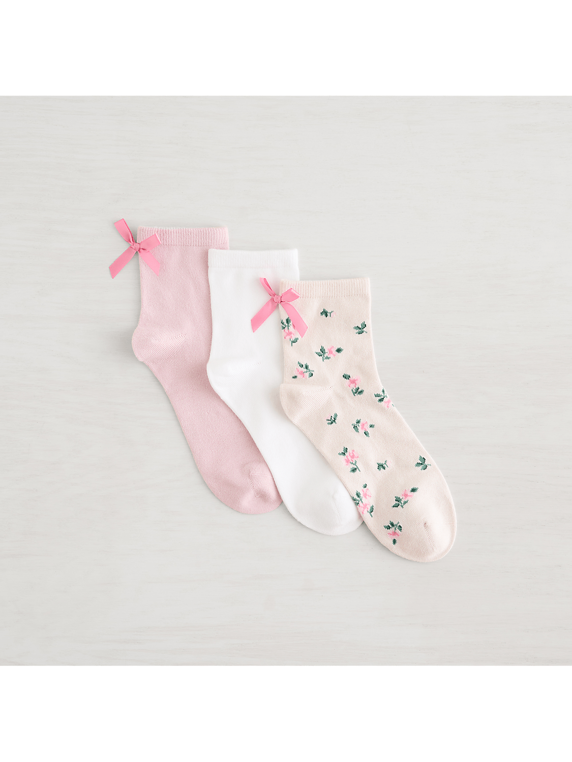 Three pairs of socks, two plain pink and white with pink bows, and one patterned with pink flowers, rest on a light wooden surface. Product Image 3