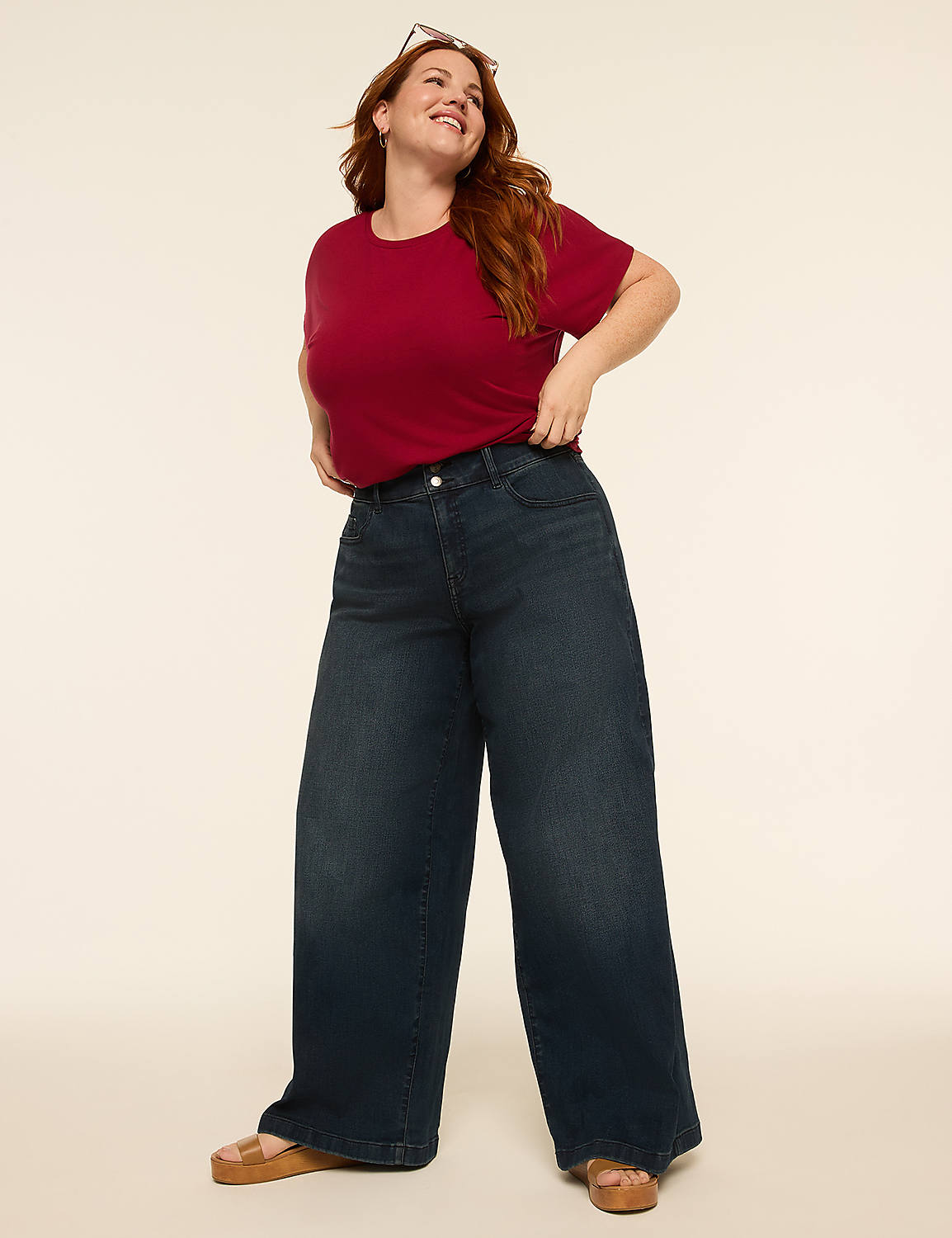 A woman with auburn hair stands in the foreground, wearing a floral print top and wide-leg jeans, holding a straw handbag, against a plain, light-colored background. Product Image 3