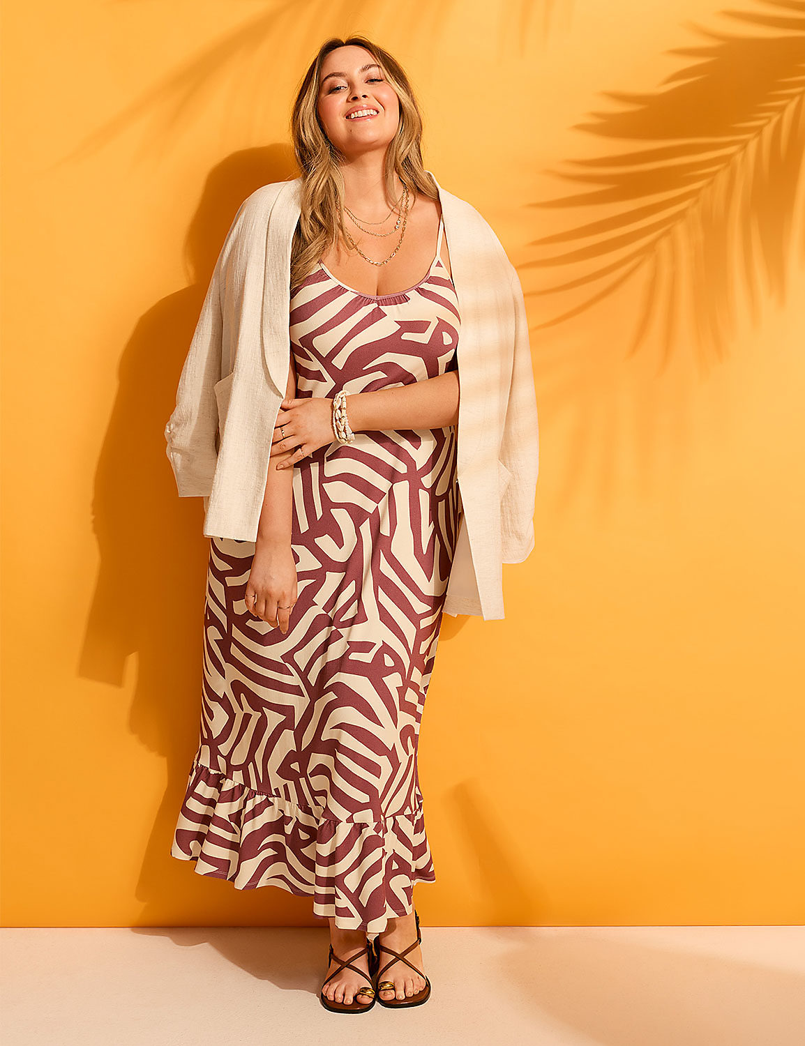 A smiling woman in a patterned dress and jacket stands against a vibrant orange background with palm leaf shadows. Product Image 5