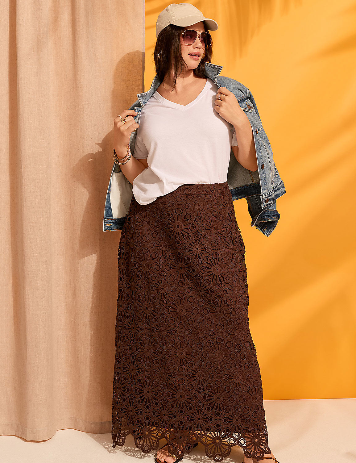 A woman in a white v-neck shirt, denim jacket, and brown lace maxi skirt stands against a backdrop of a beige curtain and an orange wall with palm leaf shadows. Product Image 4