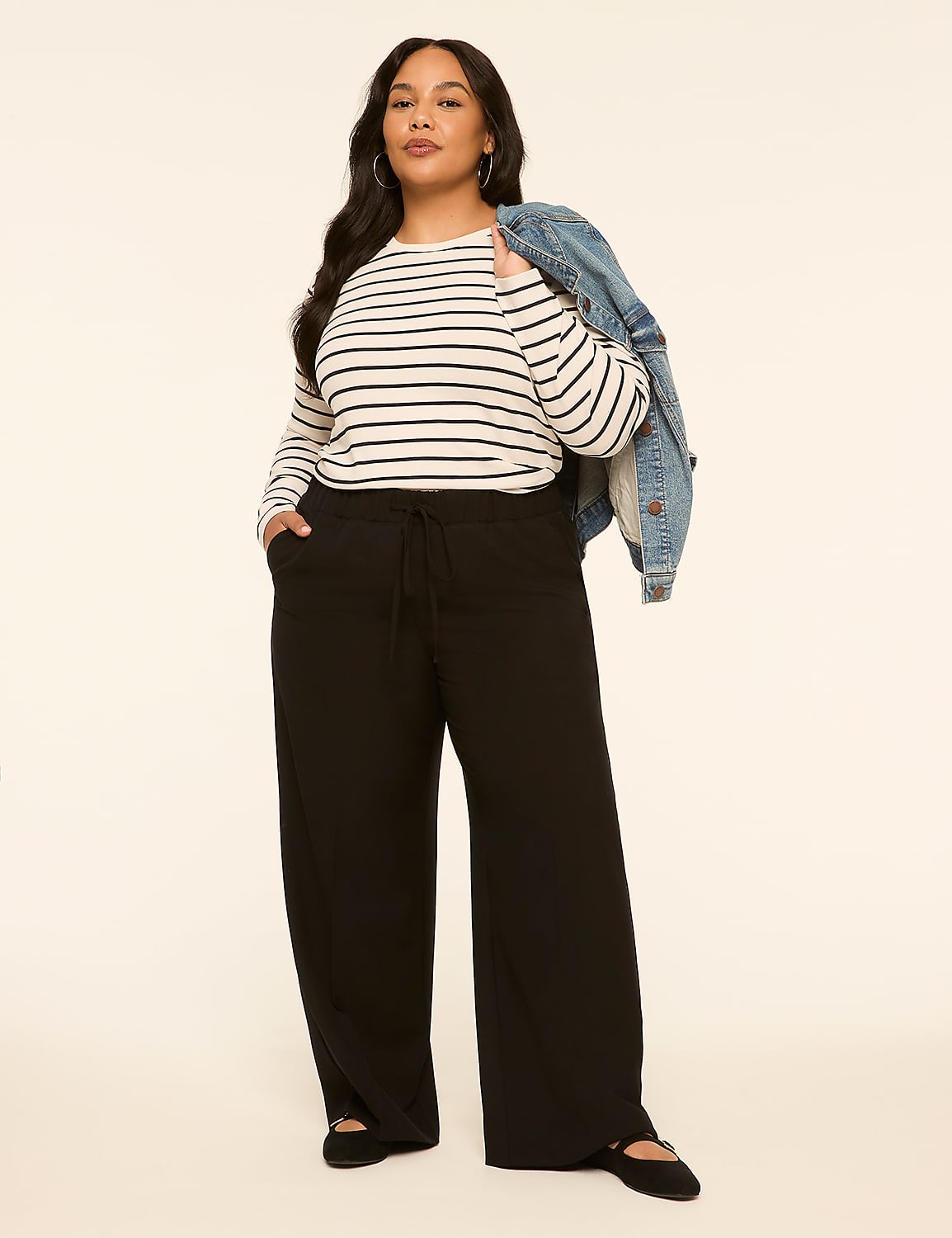 A woman in a striped shirt and black pants stands against a plain, light-colored background with a denim jacket draped over her shoulder. Product Image 3
