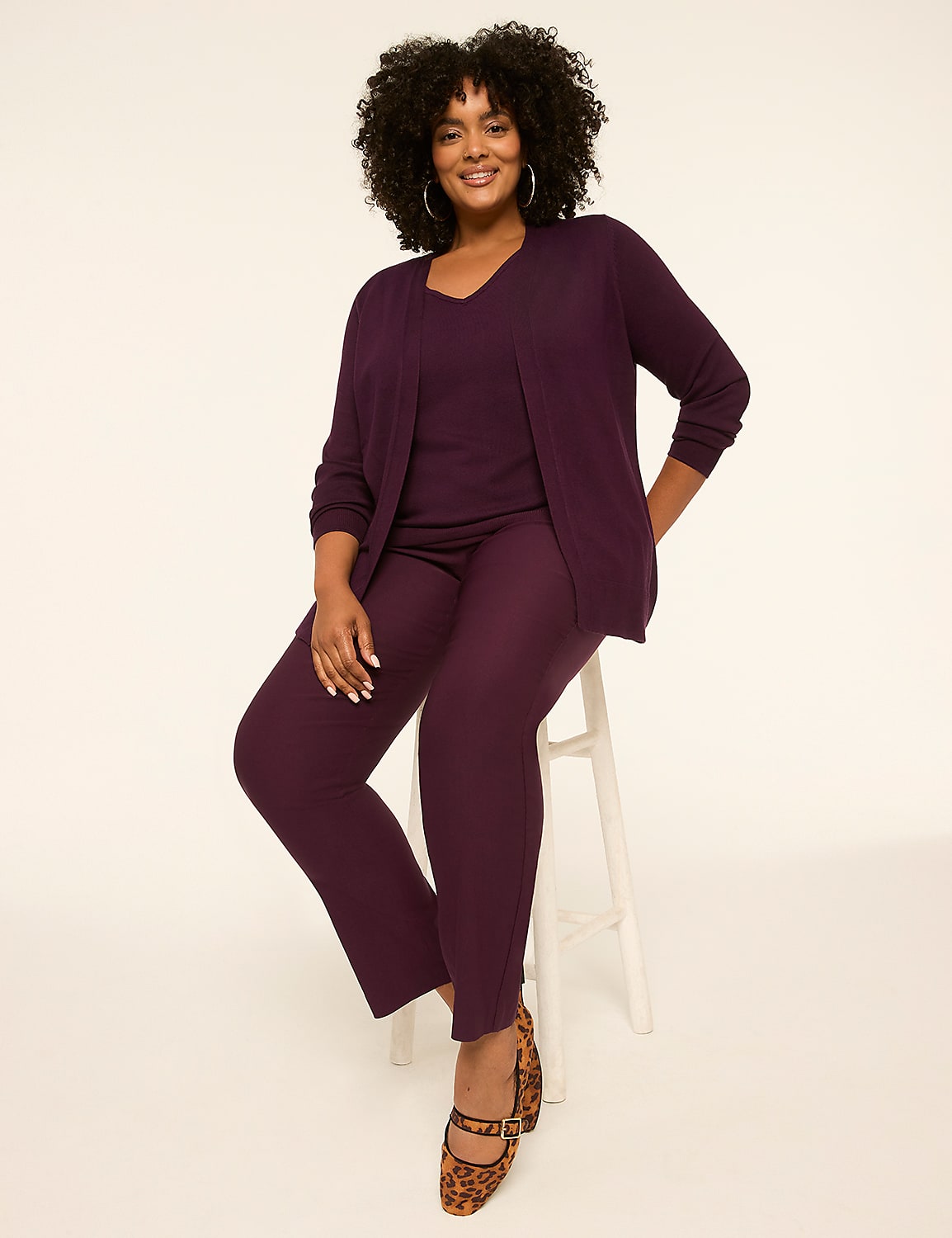 A smiling woman in a plum-colored pantsuit and cardigan sits on a white stool against a plain, light background. Product Image 3