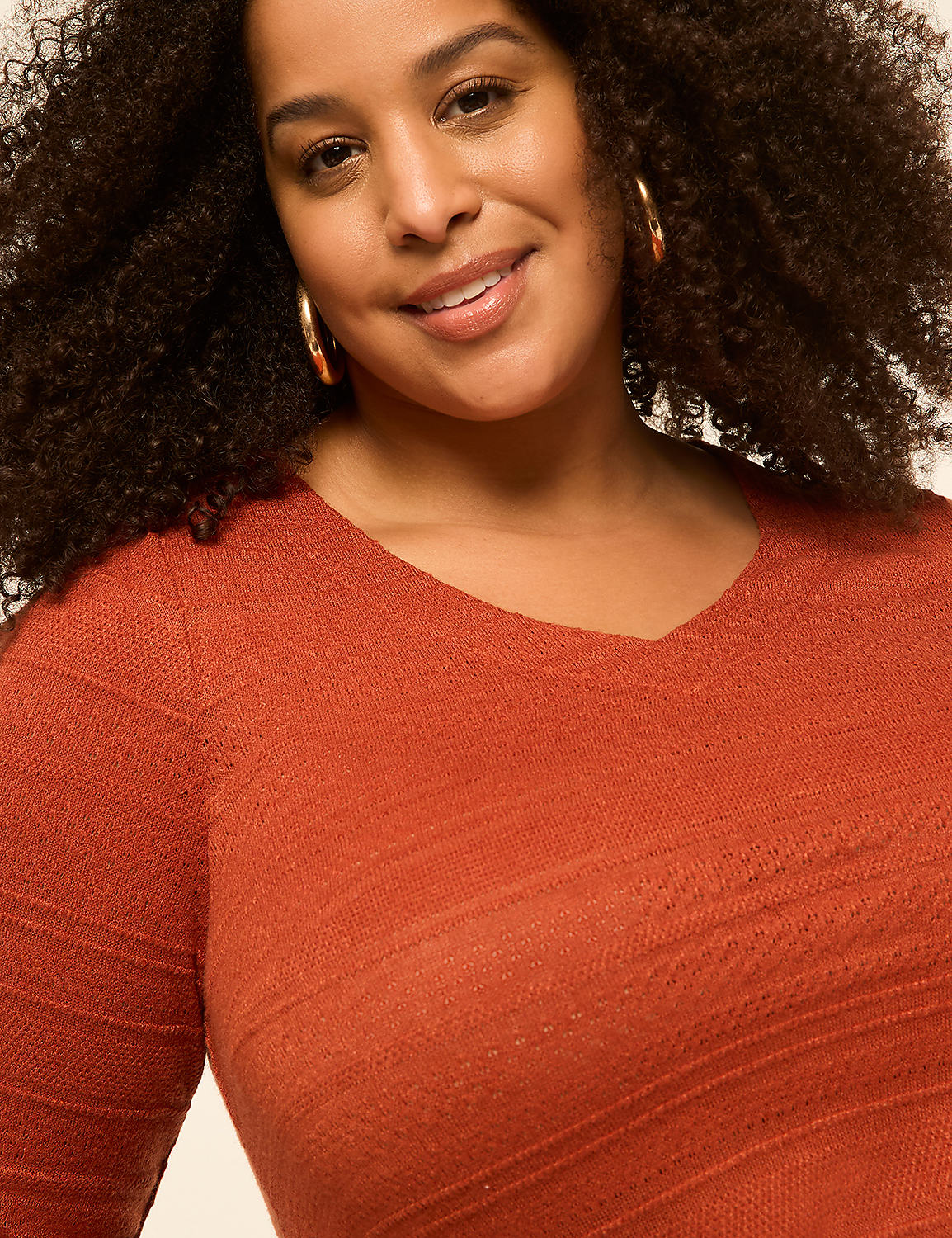 A woman with curly hair and hoop earrings smiles in the foreground, wearing an orange v-neck sweater against a plain, light background. Product Image 3