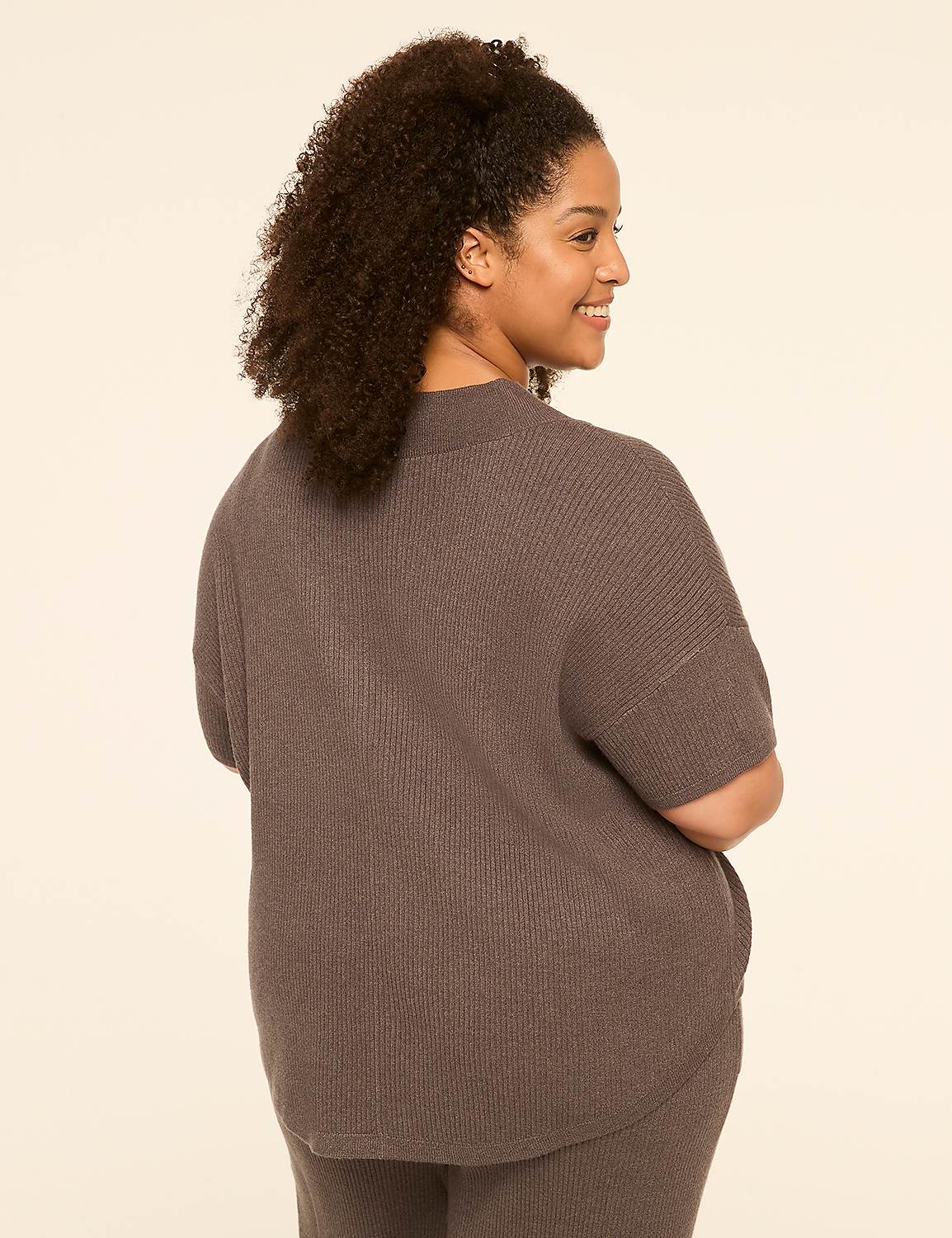 A woman with curly dark hair, wearing a gray ribbed sweater and pants, smiles over her shoulder against a plain cream background. Product Image 2