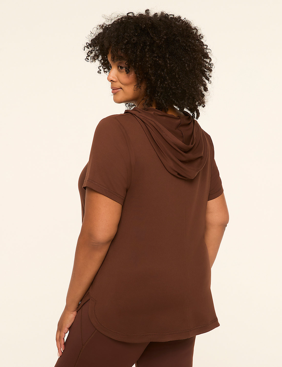 A woman with curly dark hair wears a brown short-sleeved hoodie and leggings, standing against a plain cream background. Product Image 2