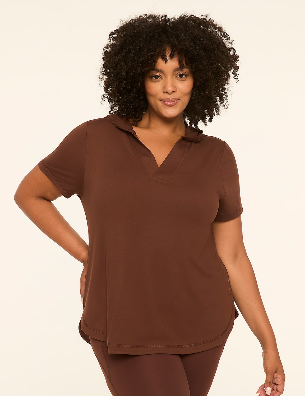 A woman with dark, curly hair and a warm smile poses in a brown polo shirt and leggings against a plain, light-colored background. Product Image 1