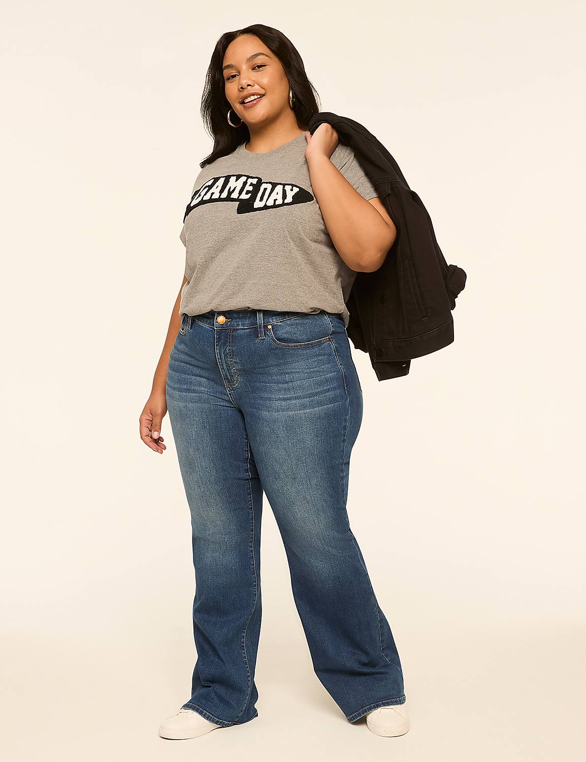 A woman in a gray "GAME DAY" t-shirt, blue jeans, and white sneakers stands against a plain, light-colored background with a white jacket draped over her shoulder. Product Image 3