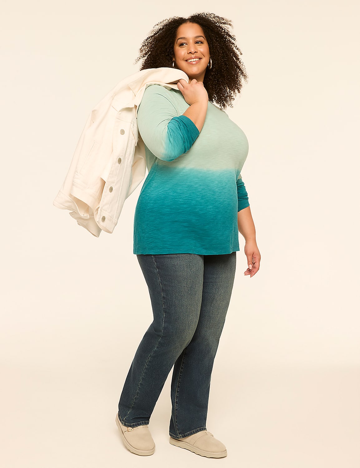 A smiling woman in a teal ombre top and jeans stands against a plain, light-colored background, with a white jacket draped over her shoulder. Product Image 3