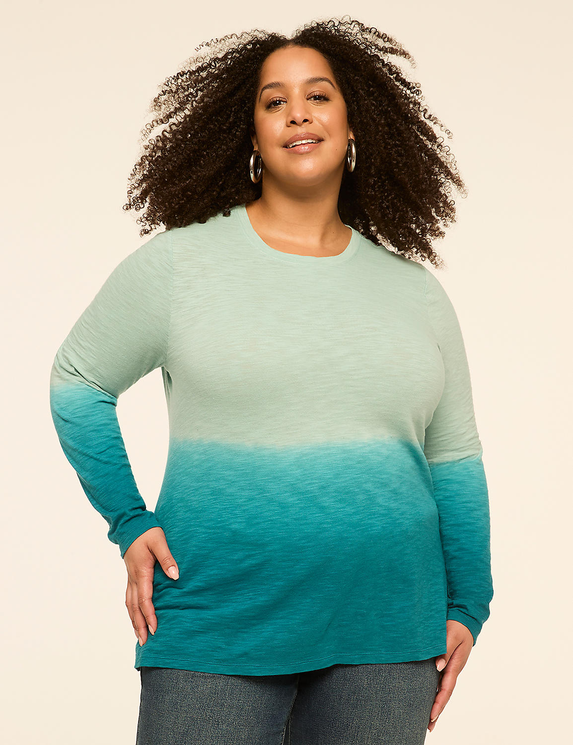 A woman with curly hair smiles at the camera, wearing a long-sleeved, ombre teal shirt and jeans against a plain, light-colored background. Product Image 1