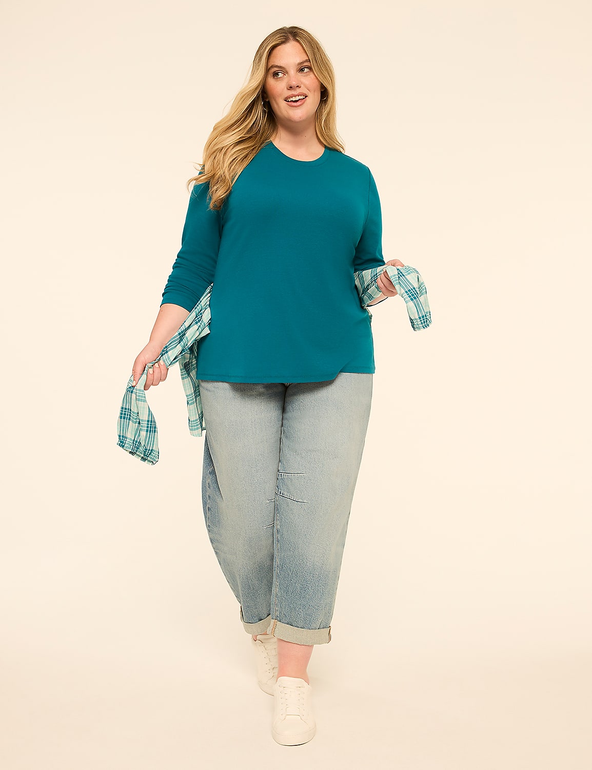 A woman in a teal long-sleeved shirt and light-wash jeans walks forward, holding a plaid shirt, against a plain cream background. Product Image 3
